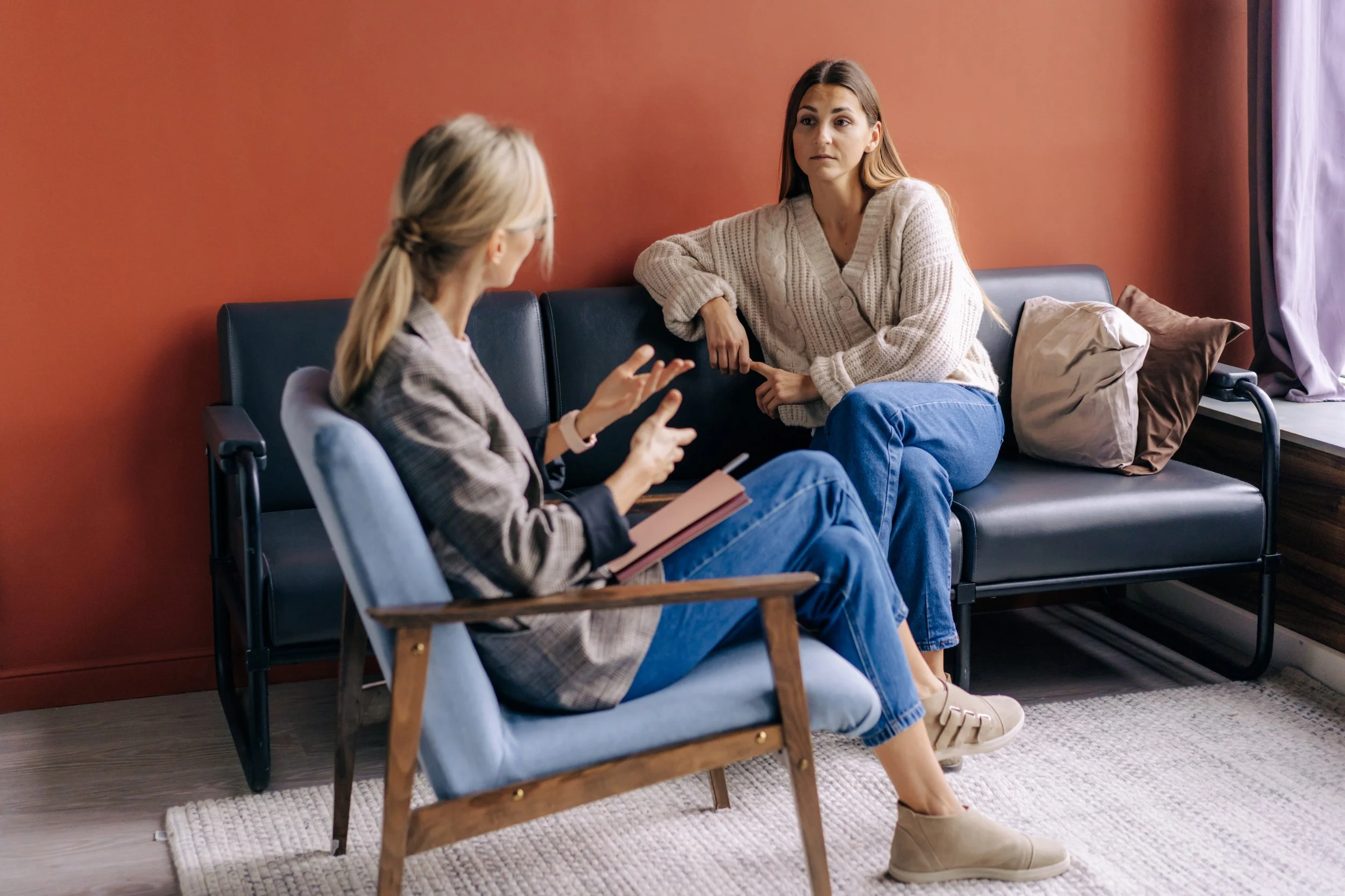 Two women are sitting and talking in a living room. One woman with glasses and blonde hair tied in a ponytail, wearing a blazer and jeans, is holding a book and gesturing. The other woman with long brown hair, wearing a beige sweater and blue jeans, is listening with her arms resting on the sofa.