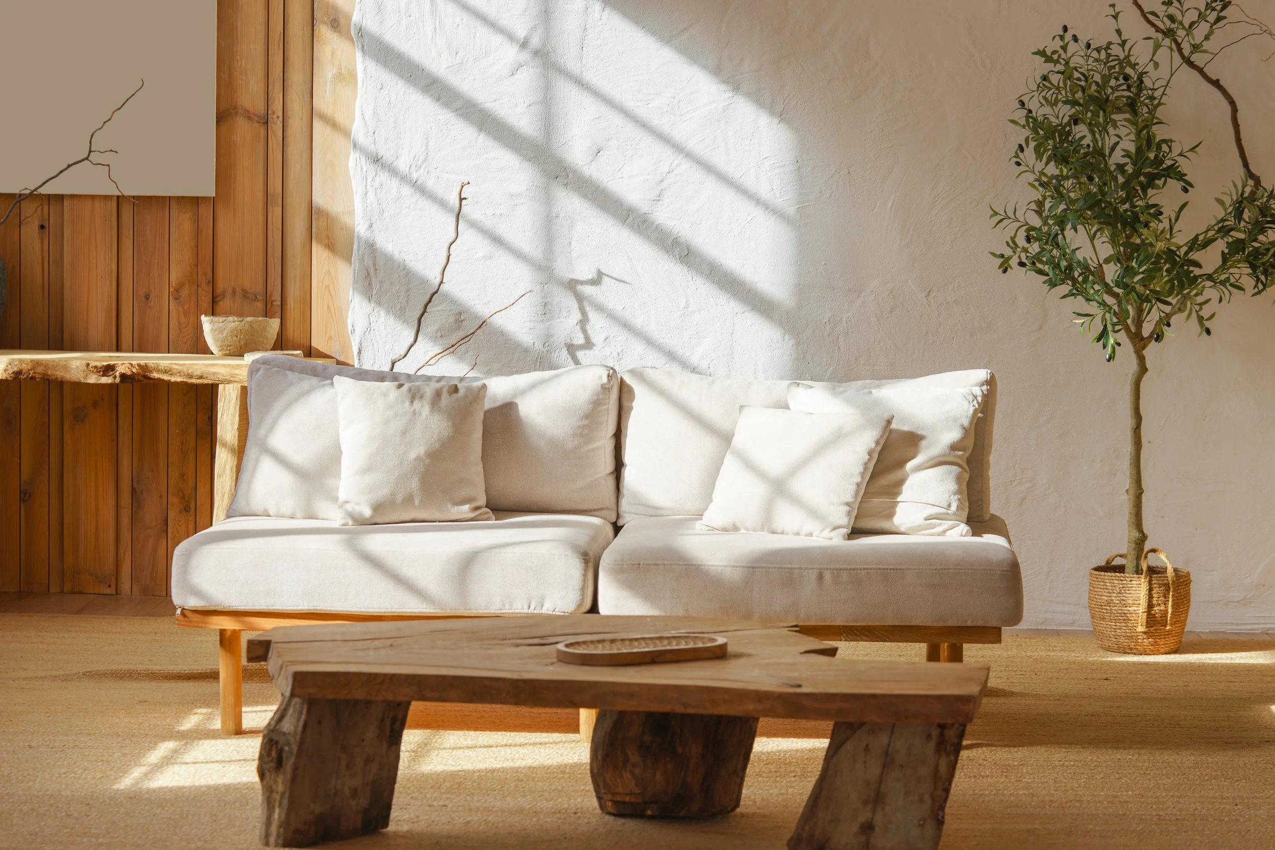 A living room with a white sofa with three matching pillows, a wooden coffee table, a potted plant in a woven basket, and a wooden accent wall. Sunlight creates shadows on the white wall.