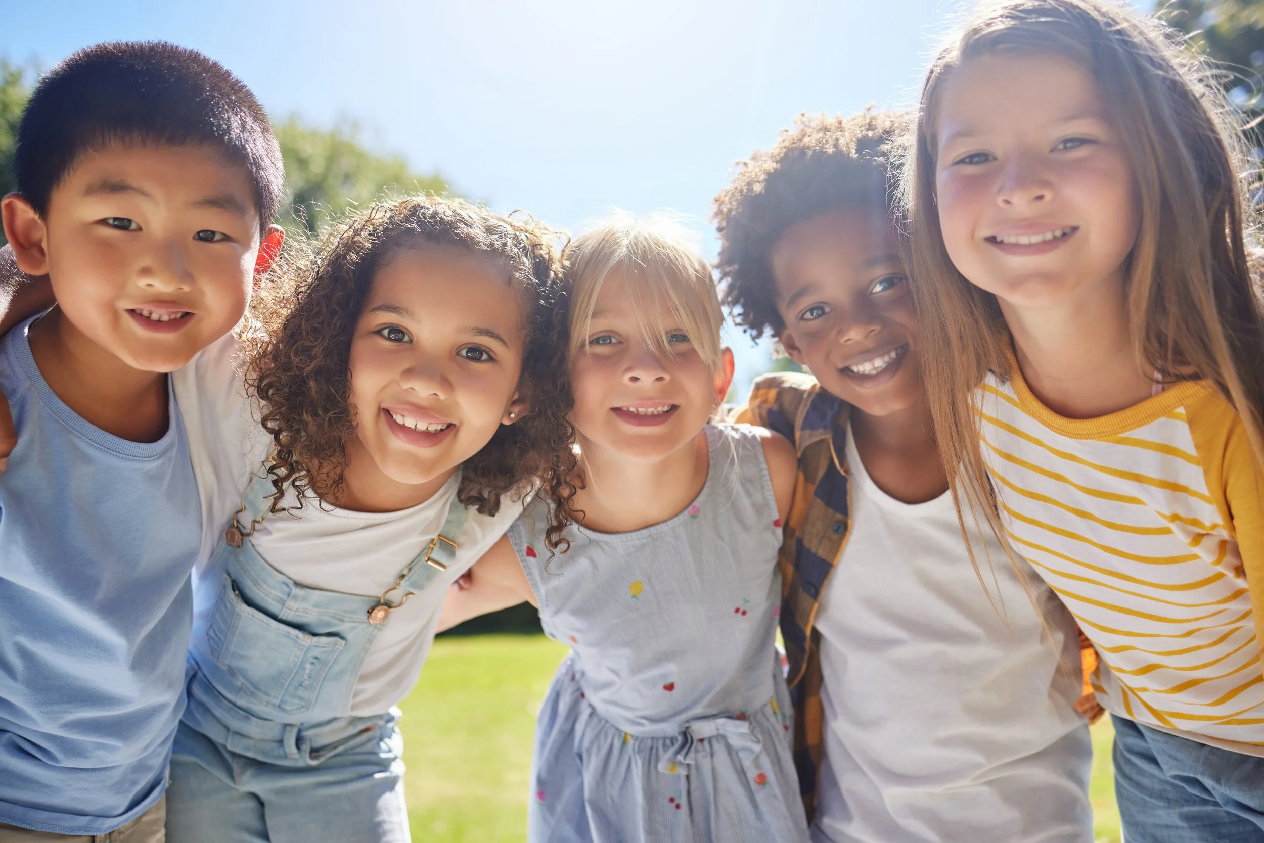 Group of six children smiling and huddled together outdoors on a sunny day