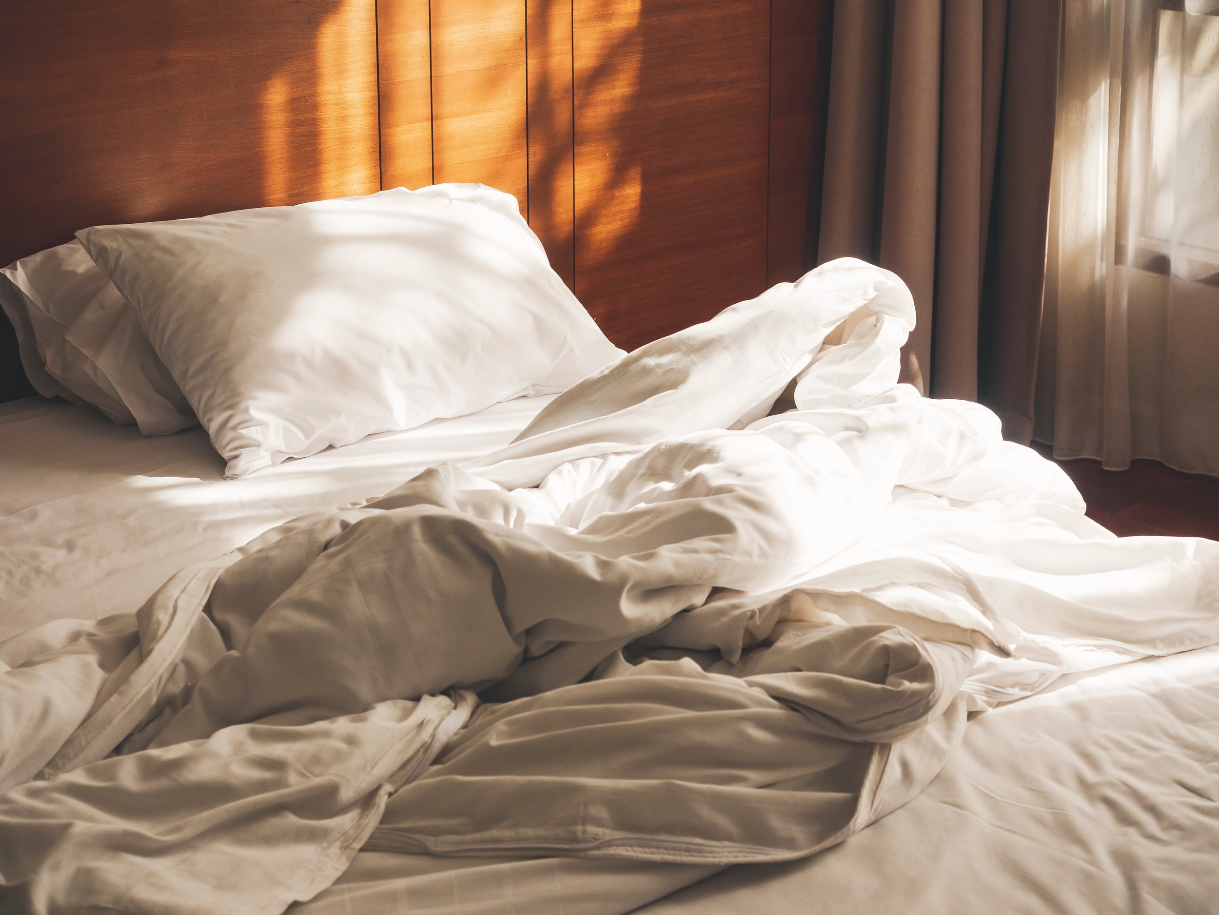 Unmade bed with white sheets and pillows, sunlight coming through a window with curtains, and a wooden headboard.