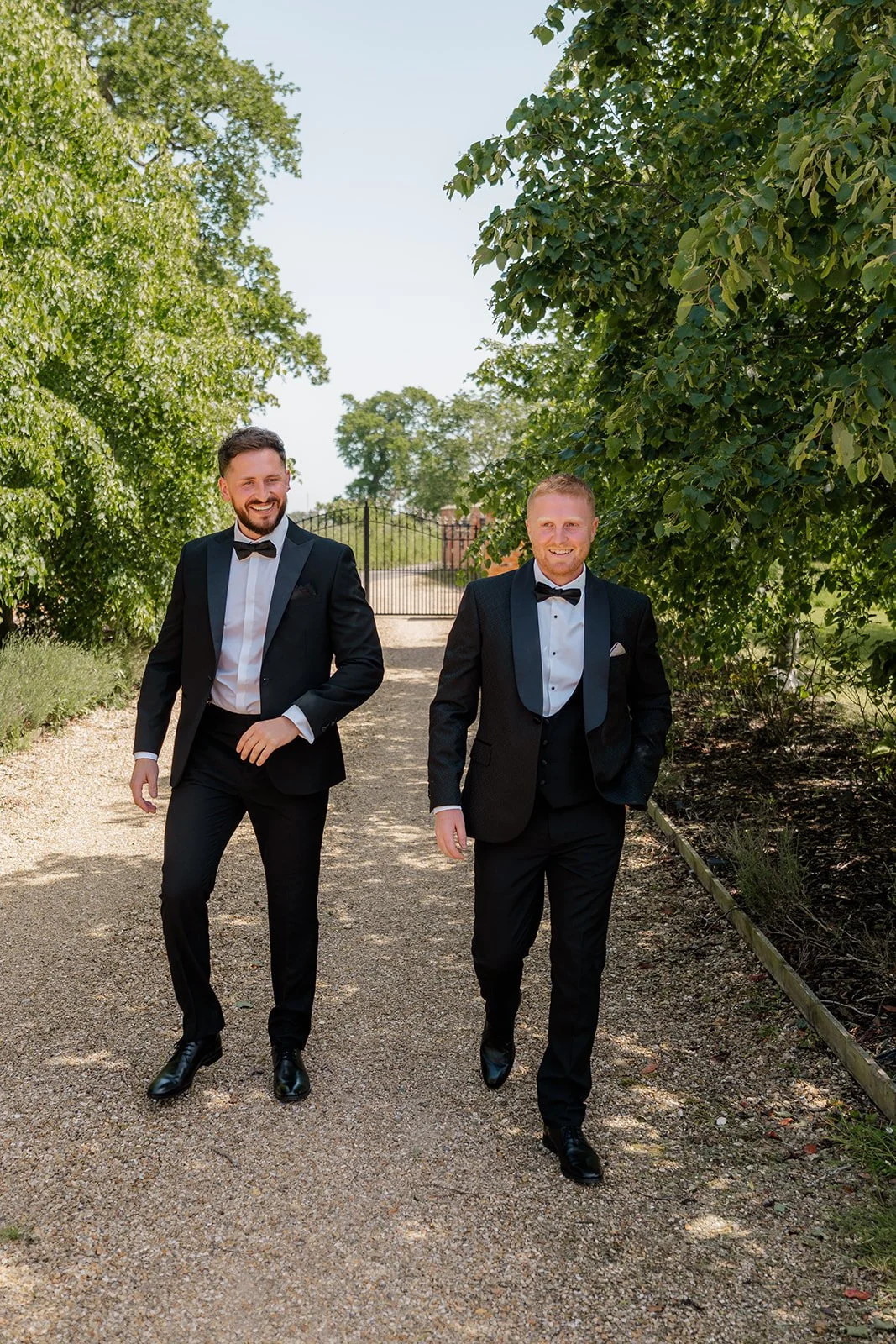 Groom and groomsman walking outside in black tuxedos beneath leafy trees at Hencote Estate in Shropshire, UK, during Lydia & Kieran's sunny wedding day, with relaxed formal style, bright greenery and a countryside venue entrance behind them.