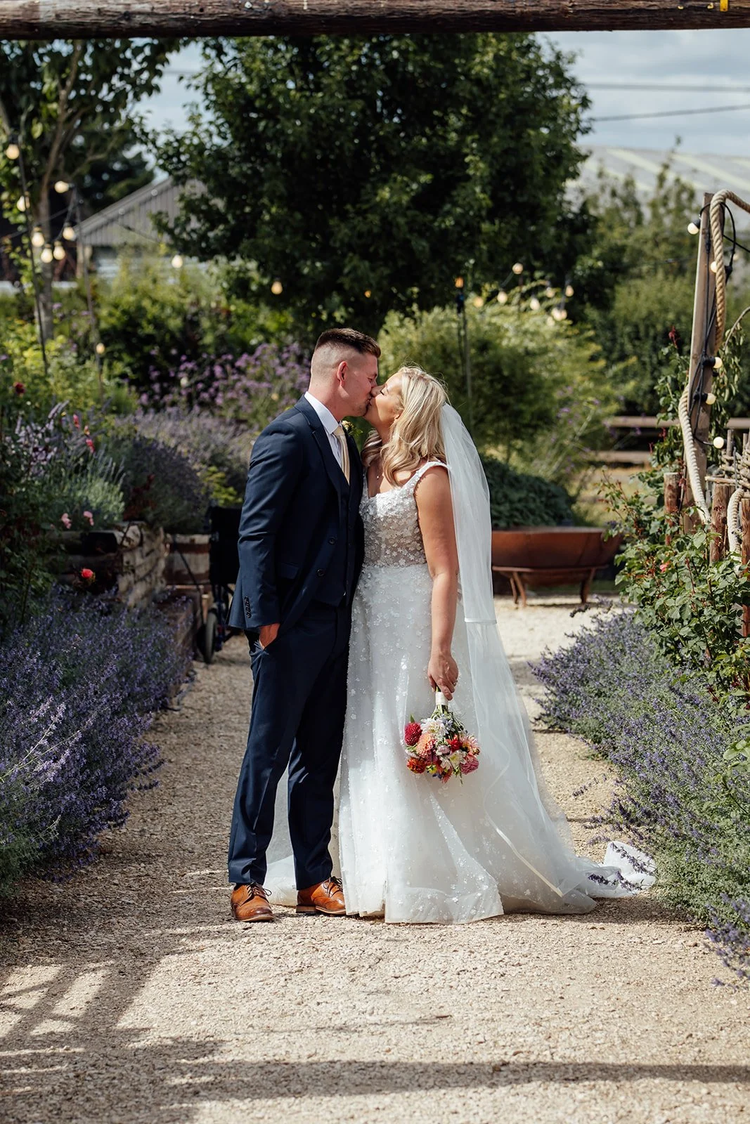 Elise and Tom sharing a kiss between lavender-lined gardens during their Bowerchalke Barn wedding portraits in Wiltshire, photographed by Just James Weddings.