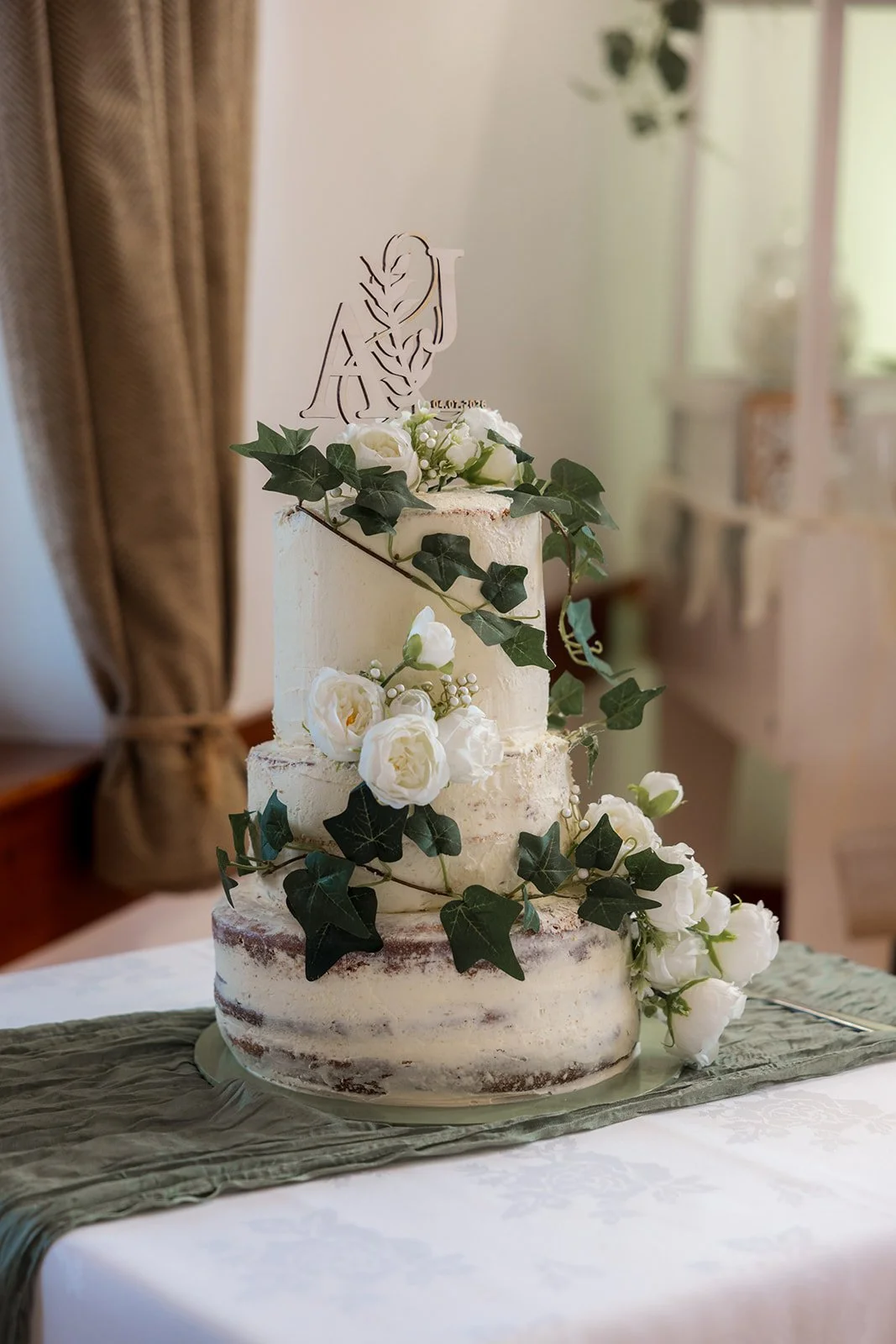 Three-tier semi-naked wedding cake decorated with white flowers, trailing ivy and an Aaron & Jack topper at Rock Village Hall in Worcestershire, photographed by Just James Weddings.