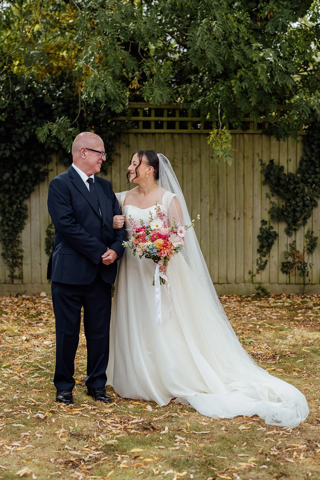 Harriet smiles with her father in a navy suit while holding her colourful bouquet before the ceremony at Dovecote Events in Oxfordshire, photographed by Just James Weddings.