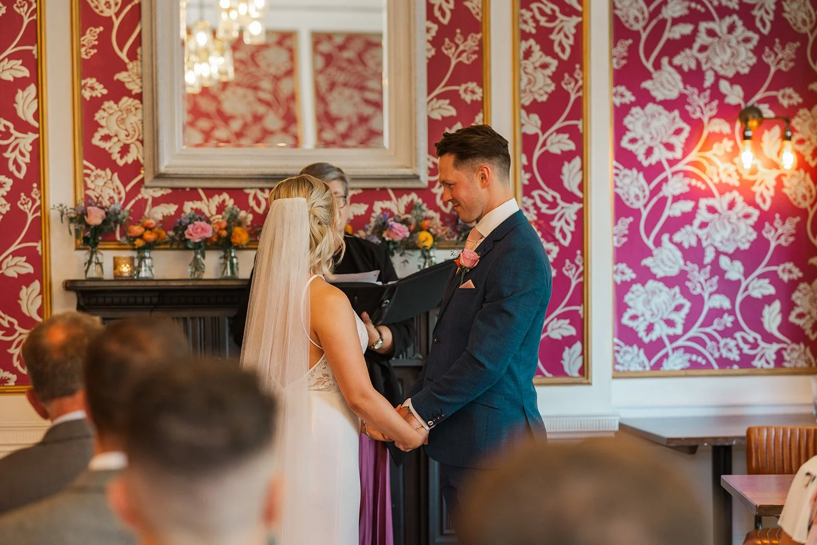 Kristy and Will hold hands during their wedding ceremony at No. 4 Clifton Village in Bristol, UK, standing together in front of the venue’s distinctive red patterned décor.
