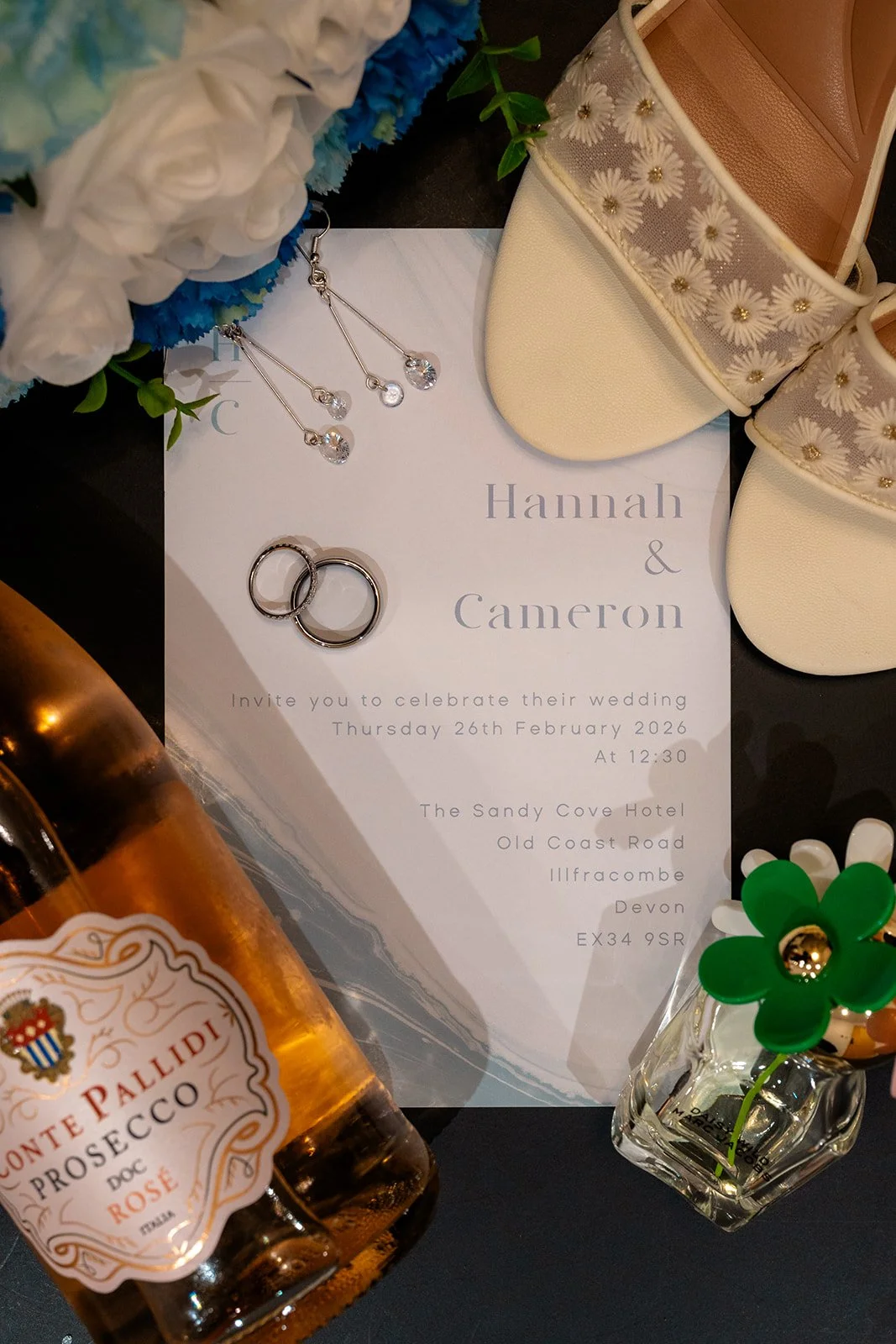 Hannah & Cameron wedding invitation flat lay with rings, bridal shoes, earrings and prosecco arranged for detail photos at Sandy Cove Hotel, North Devon. Wedding stationery and accessories photographed during Hannah & Cameron’s wedding morning.