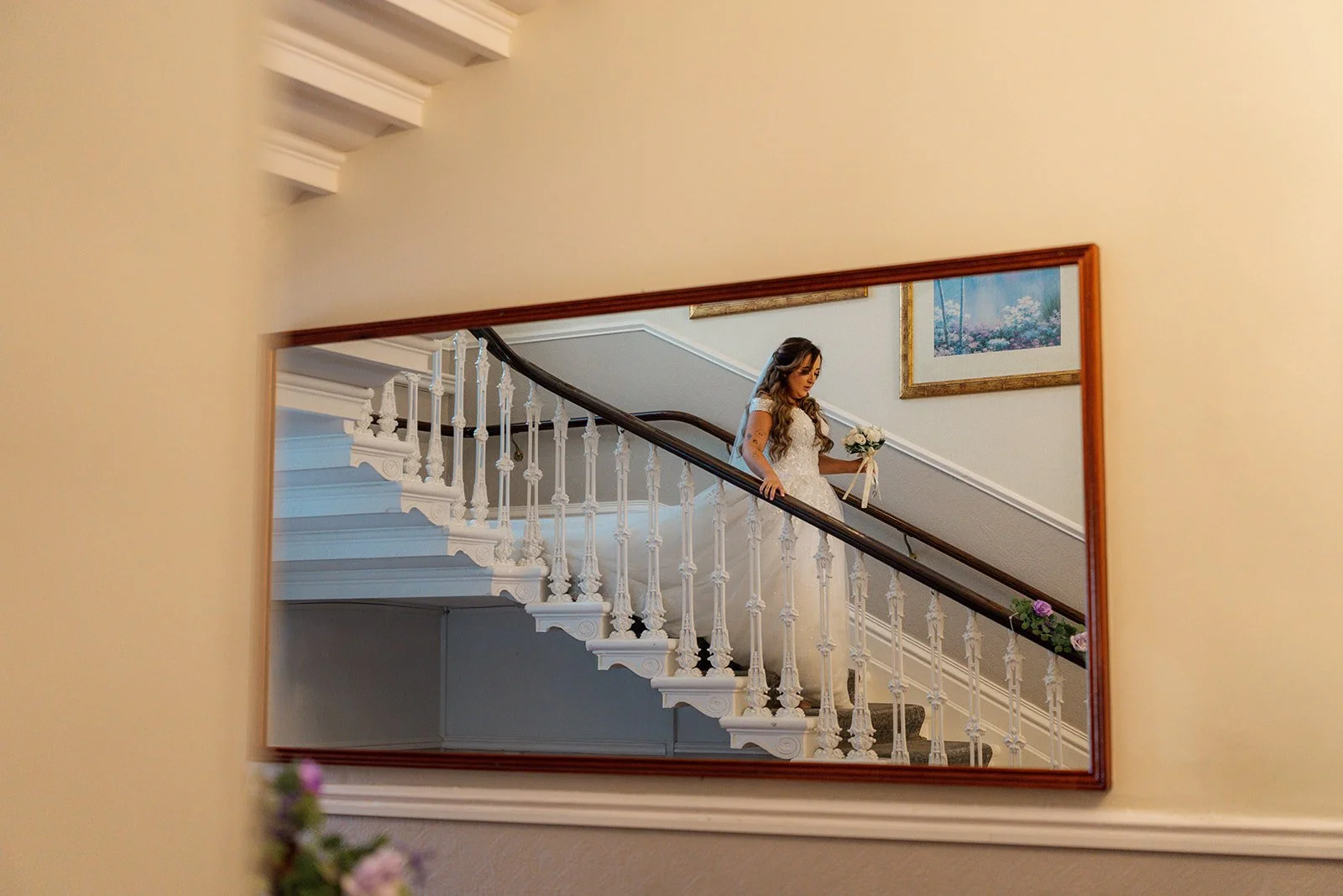 Rhiannon reflected in a wall mirror while walking down the staircase at Eastwood Park in Gloucestershire, photographed by Just James Weddings for creative bridal morning coverage.