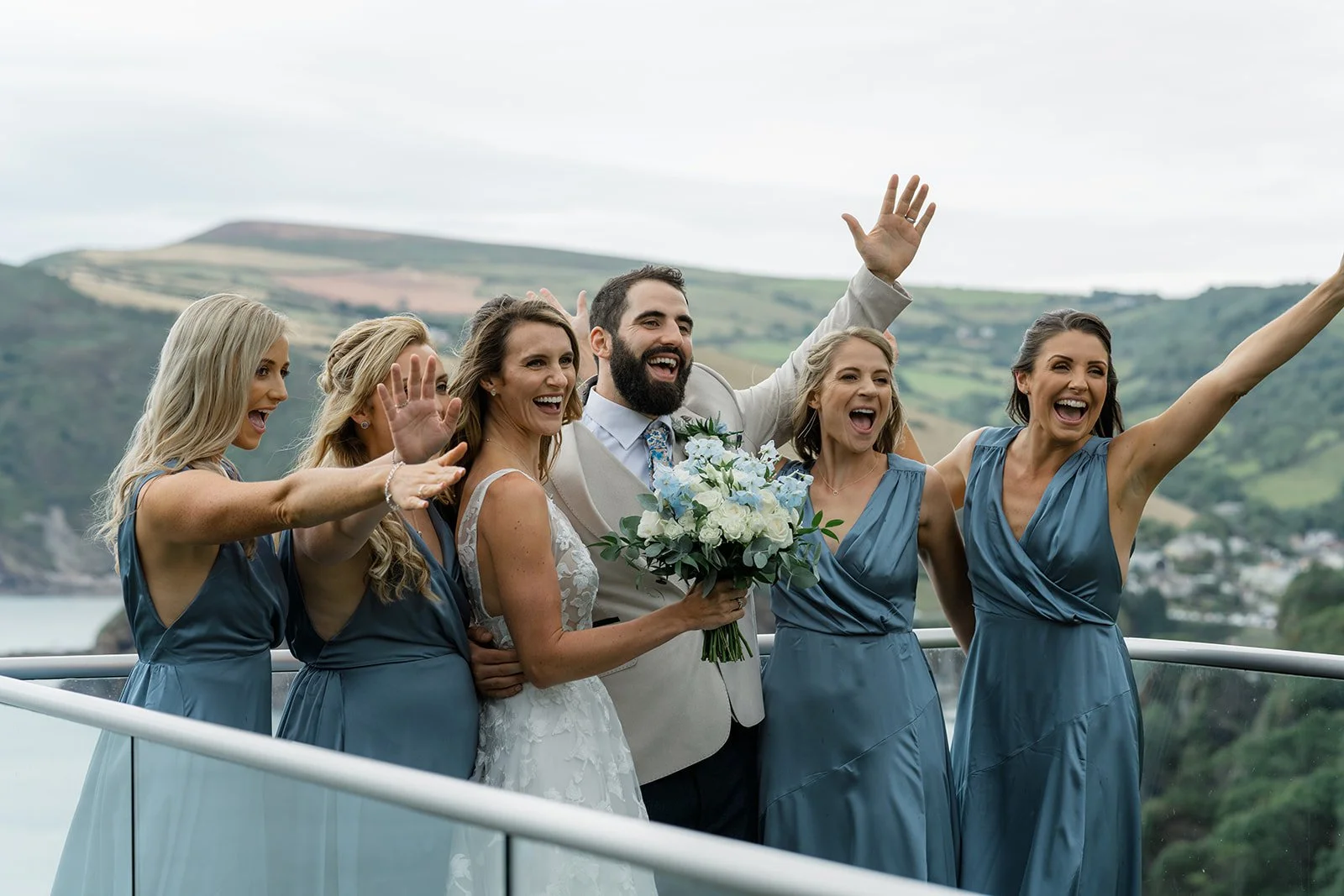 Jazz & Peter celebrating with bridesmaids on the balcony at Sandy Cove Hotel in North Devon, with sea cliffs behind them and everyone waving at the camera, photographed by Just James Weddings for fun coastal wedding party inspiration and photos.