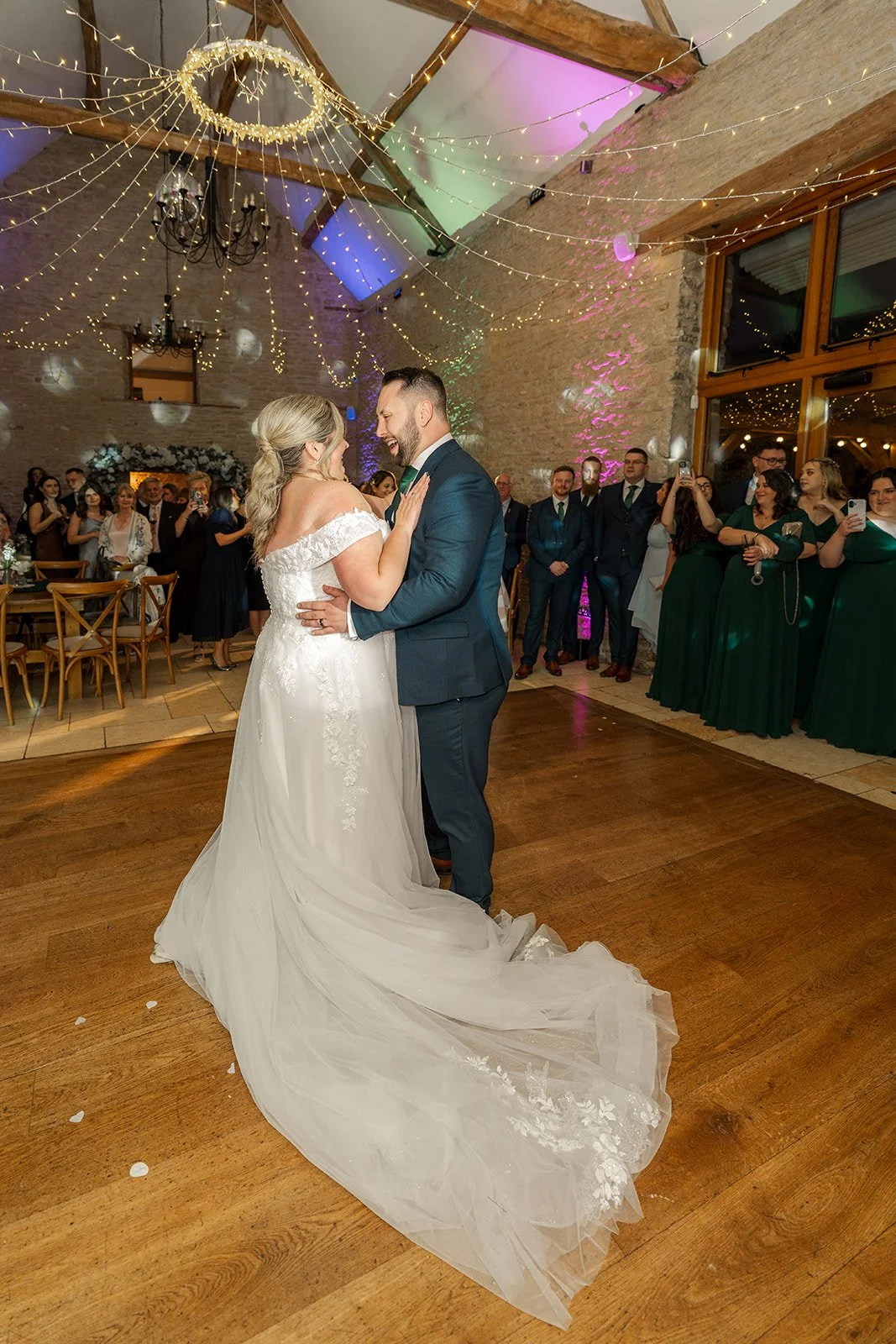 Siobhan & Nicholas share their first dance at Kingscote Barn in the Cotswolds, UK, surrounded by guests, fairy lights and colourful evening uplighting, a romantic rustic barn wedding reception moment on the wooden dance floor inside the venue.