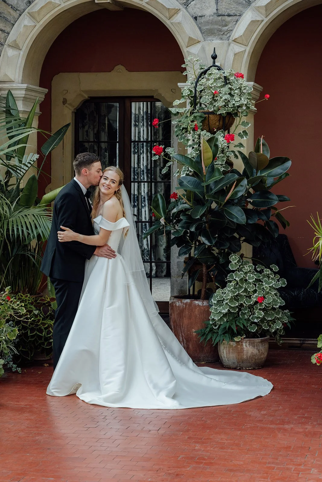 Caitlin & Kevin embracing outside Clevedon Hall in Somerset, surrounded by large potted plants and flowers, photographed by Just James Weddings.