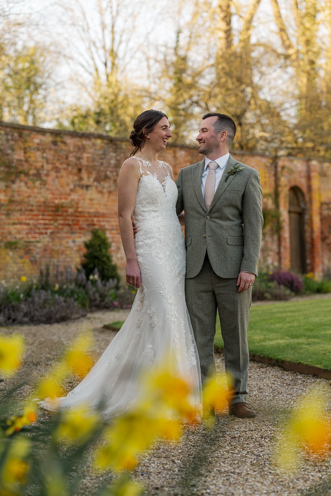 Anna and Chris stand together at Elmhay Park in Somerset, UK, smiling beside the brick garden wall as soft yellow flowers blur in the foreground. A romantic wedding portrait from Anna & Chris’s day, full of warmth, elegance, and spring colour.