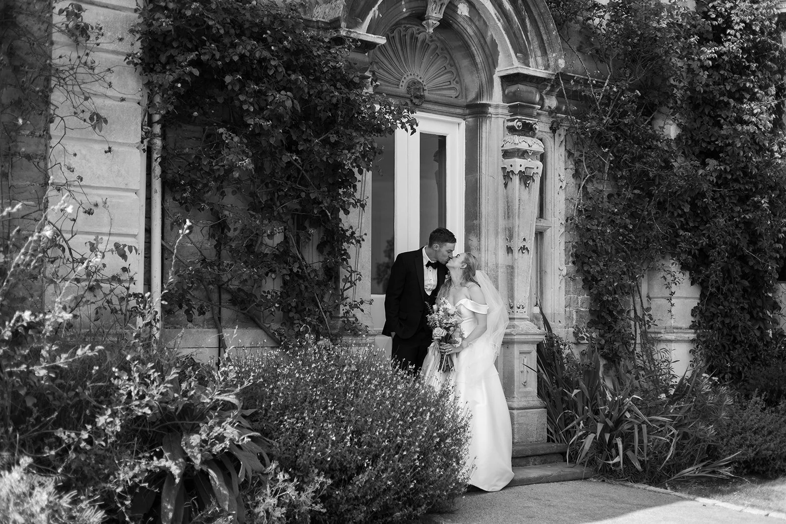 Black and white portrait of Caitlin & Kevin standing together outside an ornate doorway at Clevedon Hall in Somerset, surrounded by garden greenery, photographed by Just James Weddings.