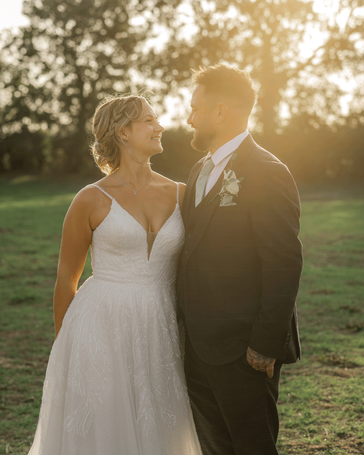 Harlie & Dave standing together in a grassy field in Glastonbury, somerset, on their wedding day. The bride smiles up at the groom as golden sunlight filters through the trees behind them, golden light across their faces and wedding outfits.