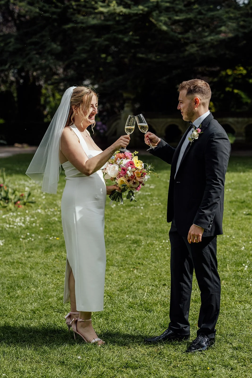 Bryony & Max toast with champagne on the lawn at Insole Court in Cardiff, smiling in the sunshine with Bryony holding her colourful bouquet as they enjoy a relaxed newlywed moment in the gardens after their intimate ceremony together nearby.