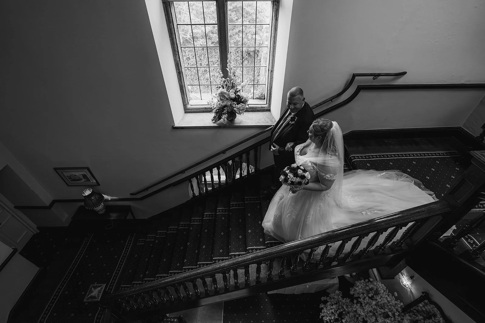 Becky and dad share a quiet moment on the grand staircase beneath a window at Clearwell Castle, Gloucestershire, UK. Bride with veil and bouquet beside groom in suit, captured in classic black and white Clearwell Castle wedding photography inside.