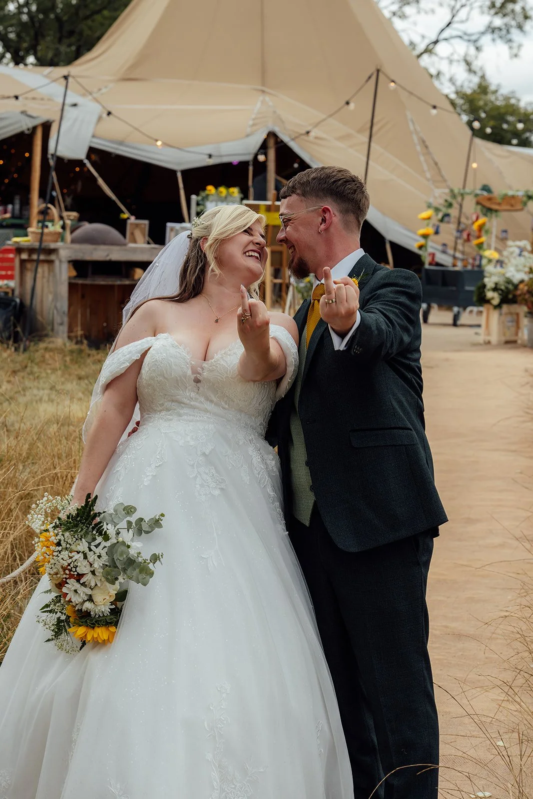 Lucy and Matthew laughing together and showing their wedding rings outside the tipi reception at Curated Canvas at Noah’s Pond in Somerset, photographed by Just James Weddings.