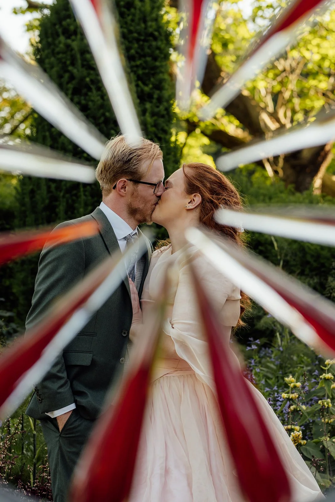 Charlotte & Jack kiss in the gardens at Bishop’s Palace in Somerset, UK, framed by blurred red and white bunting, with the bride in her pink dress and the groom in a green suit during relaxed couple portraits after their sunny wedding ceremony.