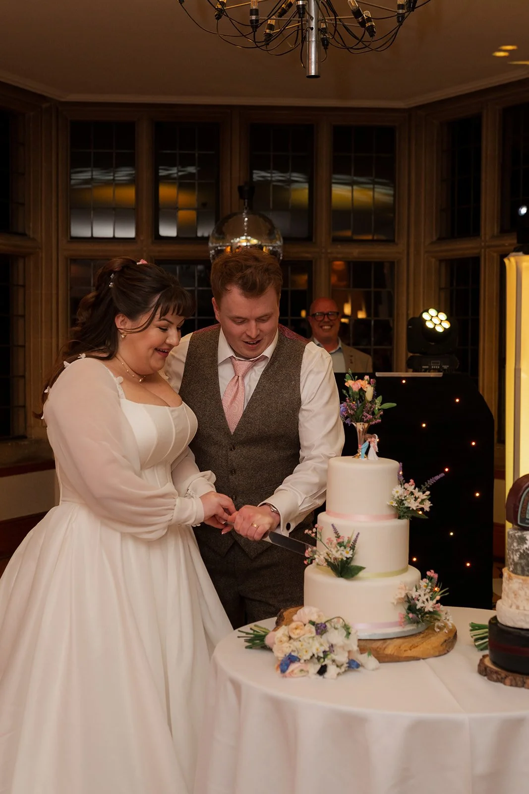 Emily & Josh cut their wedding cake together at Coombe Lodge Blagdon, Somerset, UK, beside a three-tier floral cake in the evening reception room. Indoor wedding photo with warm lighting, wood-panelled windows and guests watching behind them.