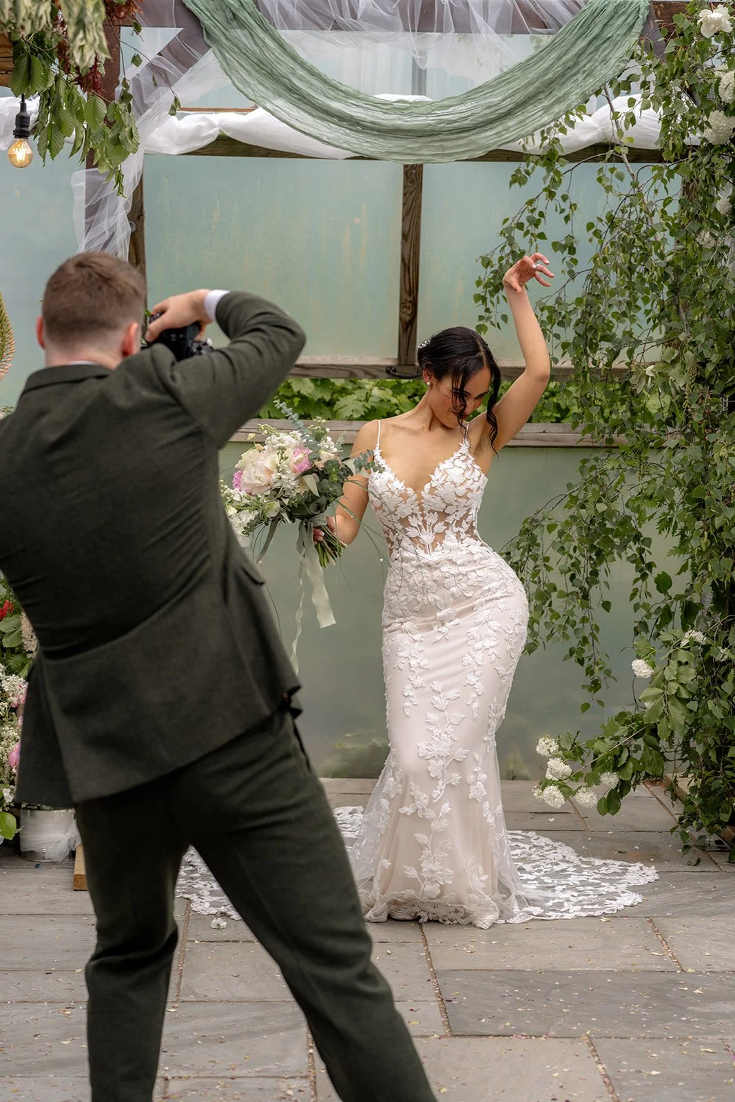 Jaye & Josh during playful wedding portraits at Farrington's Farm in Bristol, with the bride posing for the groom's camera in her lace dress, surrounded by hanging greenery, soft draped fabric and the relaxed poly tunnel ceremony setting too.
