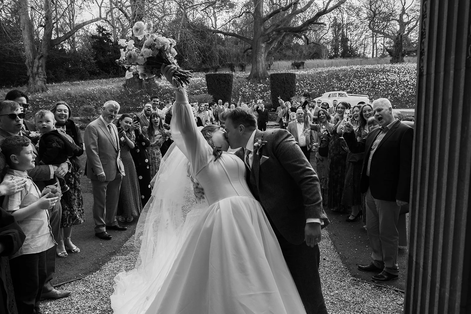 Black and white photo of Emily and Josh sharing a kiss beneath a raised bouquet after their wedding ceremony at Coombe Lodge in Somerset, UK, surrounded by cheering guests.