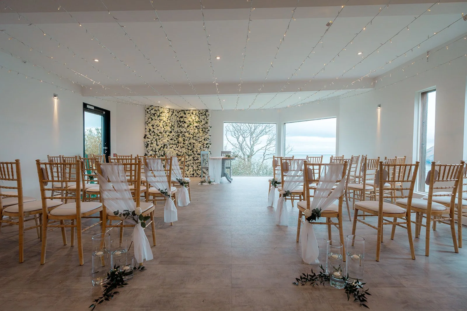 Indoor ceremony room at Sandy Cove Hotel in North Devon for Daisy & Andrew’s wedding, with gold chairs, white aisle drapes, fairy lights and sea views, photographed by Just James Weddings for relaxed coastal wedding inspiration and venue ideas.