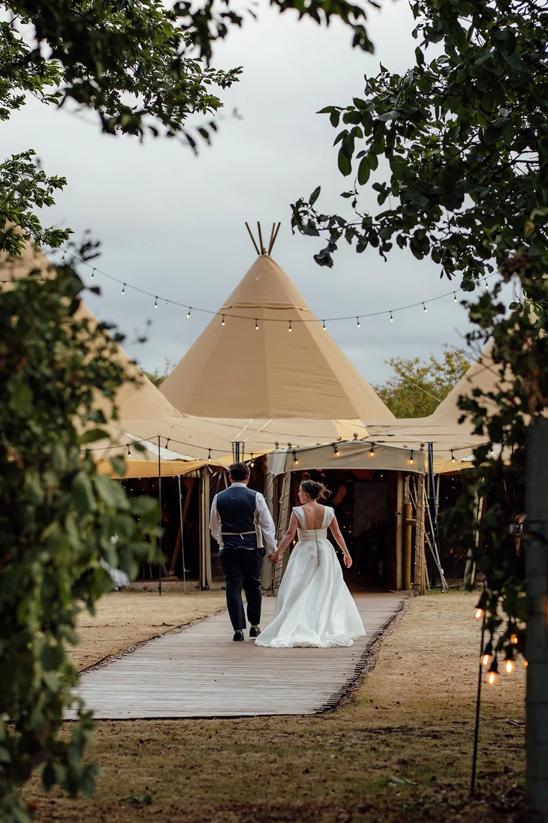 Harriet & Simon walk hand in hand towards the tipi reception beneath festoon lights at Dovecote Events in Oxfordshire, photographed by Just James Weddings.