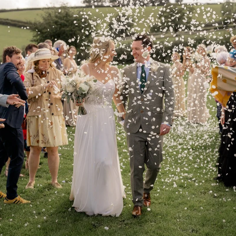 Samantha & Mike walking through a shower of white confetti after their ceremony at The Hawke Barn, devon. Guests line the grass on both sides as the bride and groom smile at each other, holding hands with rolling countryside behind them.