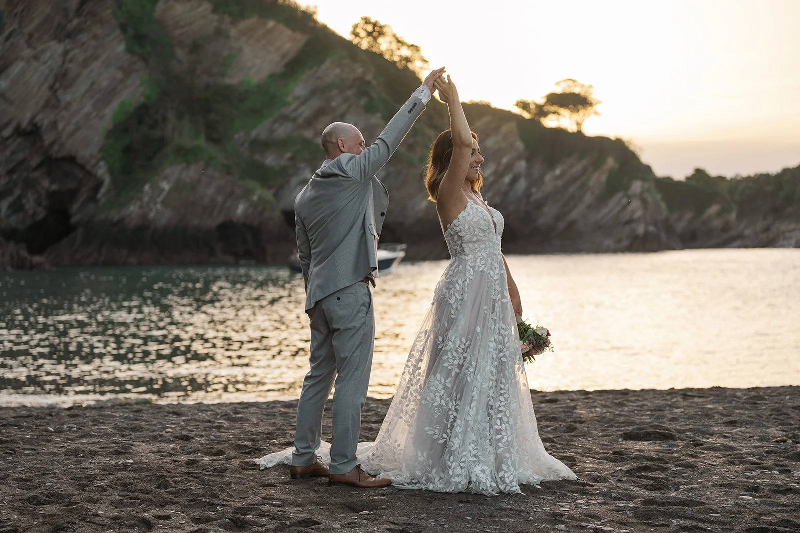 Toya & Simon dancing on the beach at sunset at Sandy Cove Hotel in North Devon, with cliffs and sea behind them as the groom twirls the bride, photographed by Just James Weddings for relaxed coastal wedding portraits in golden evening light.