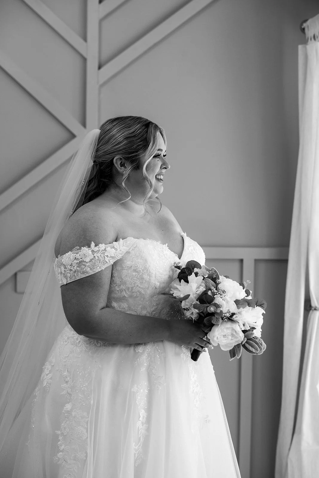 Black and white bridal portrait of Siobhan holding her bouquet and smiling by the window at Kingscote Barn in the Cotswolds, UK, during Siobhan & Nicholas' wedding morning, bridal prep photography in a rustic barn setting before the ceremony.