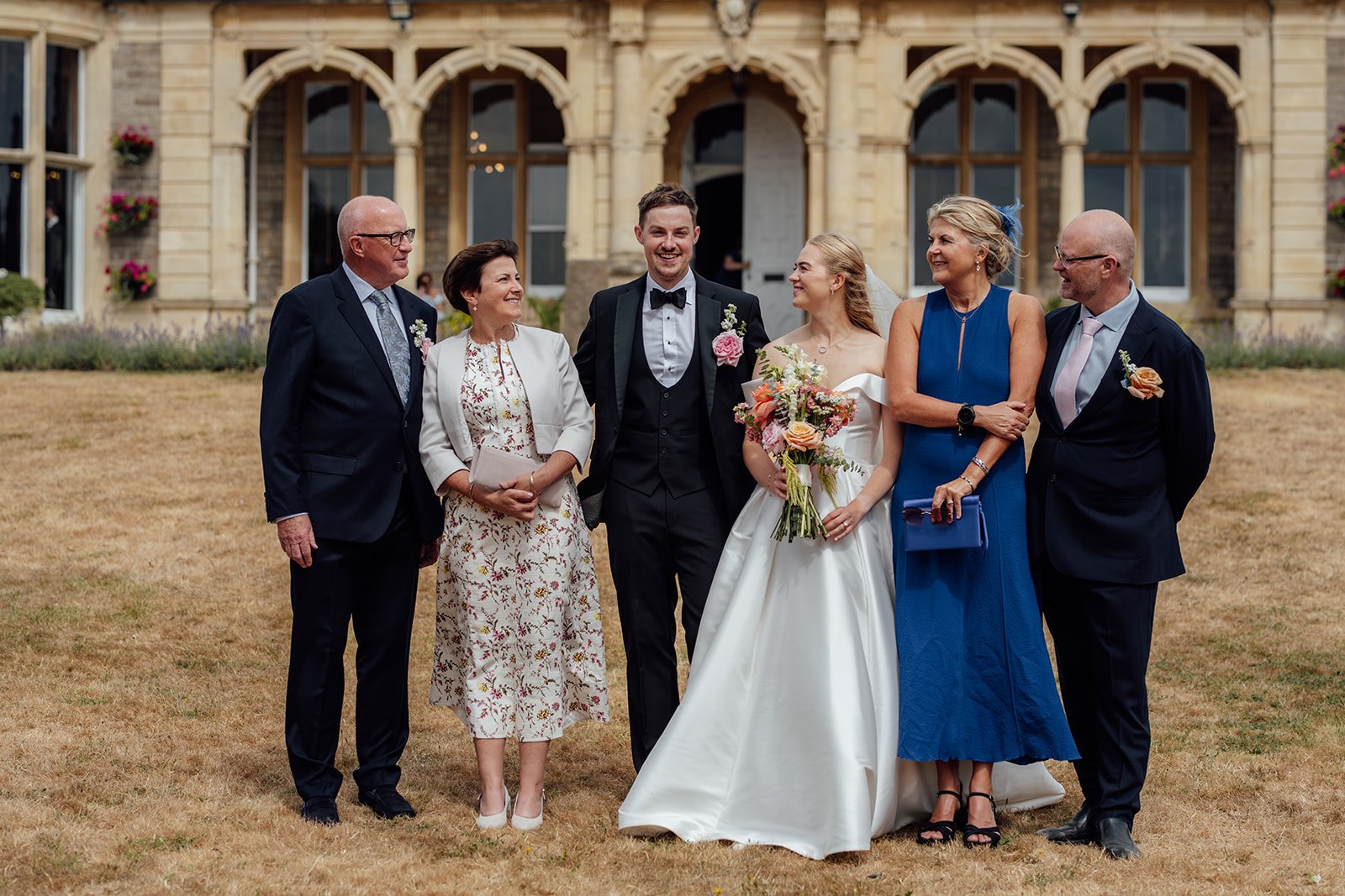 Caitlin and Kevin posing with their parents for a family wedding portrait on the lawn at Clevedon Hall in Somerset, with the historic venue in the background.