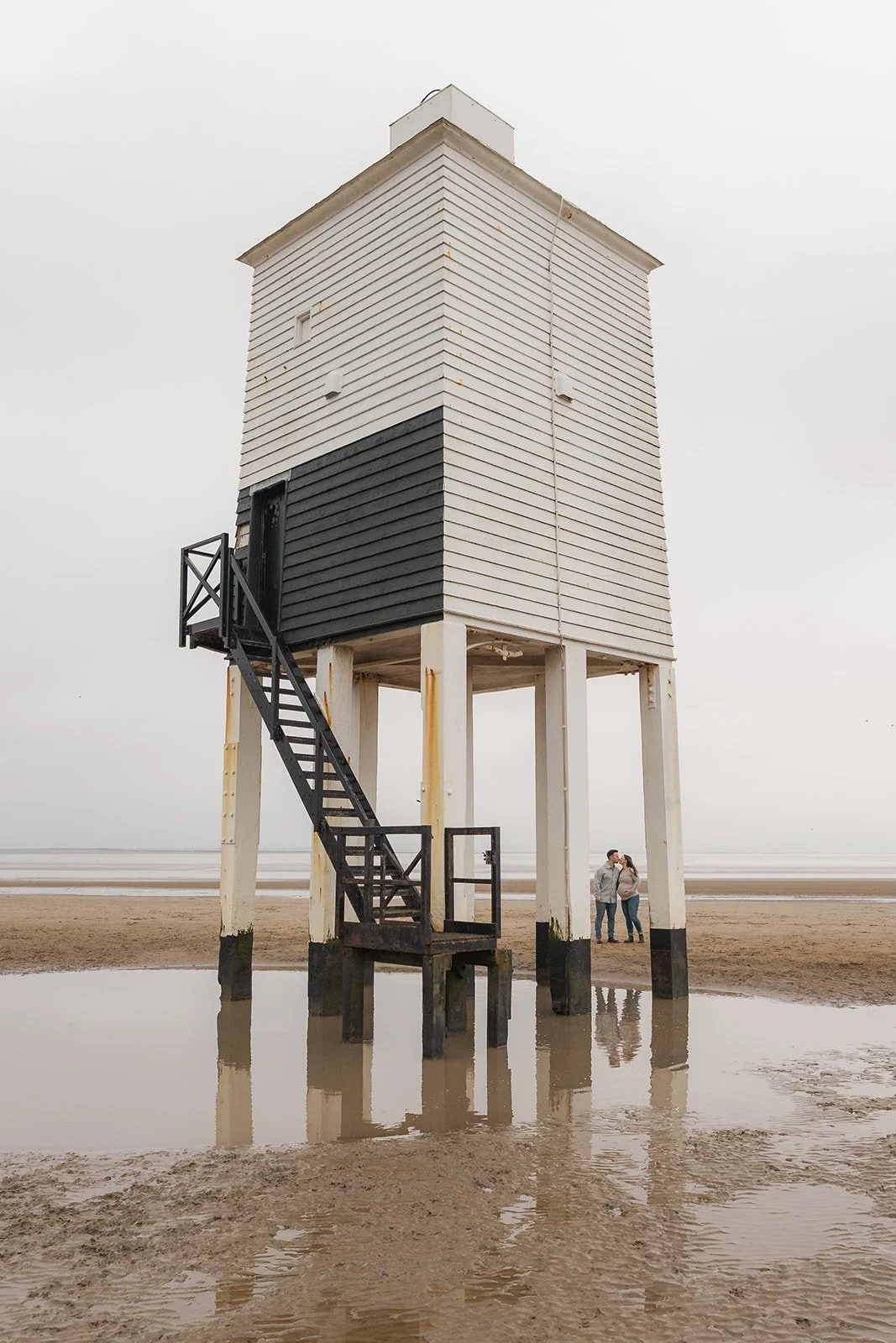Emma & Dan stand together during their engagement and maternity shoot beside a tall white lighthouse on the beach. Reflections in the wet sand and the wide open shoreline create a calm coastal scene with a quiet, intimate and documentary feel.