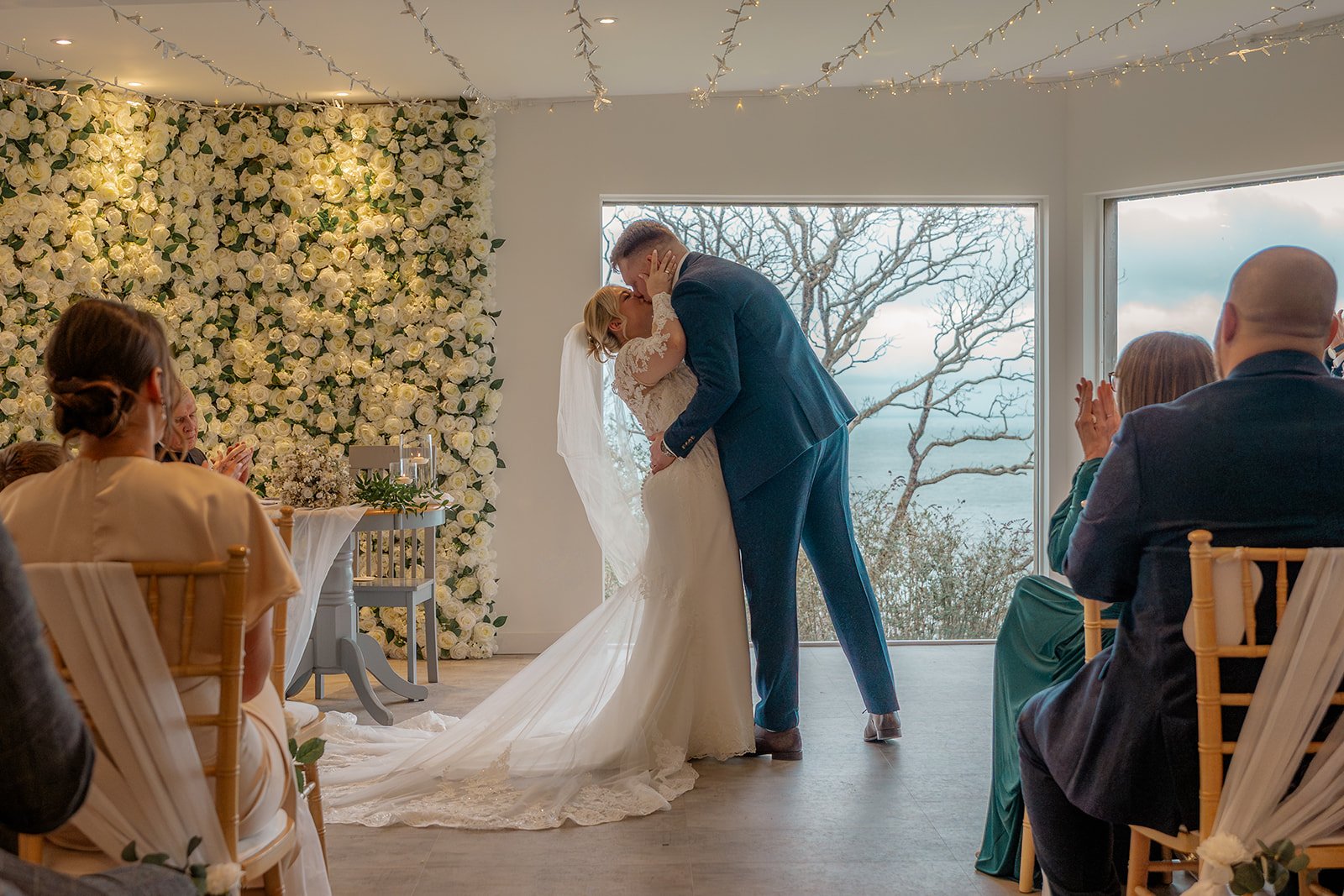 Andrew & Daisy sharing their first kiss as newlyweds during their ceremony at Sandy Cove Hotel in Devon, framed by a white floral backdrop and coastal sea views, capturing romantic indoor wedding vows at this stunning seaside venue.