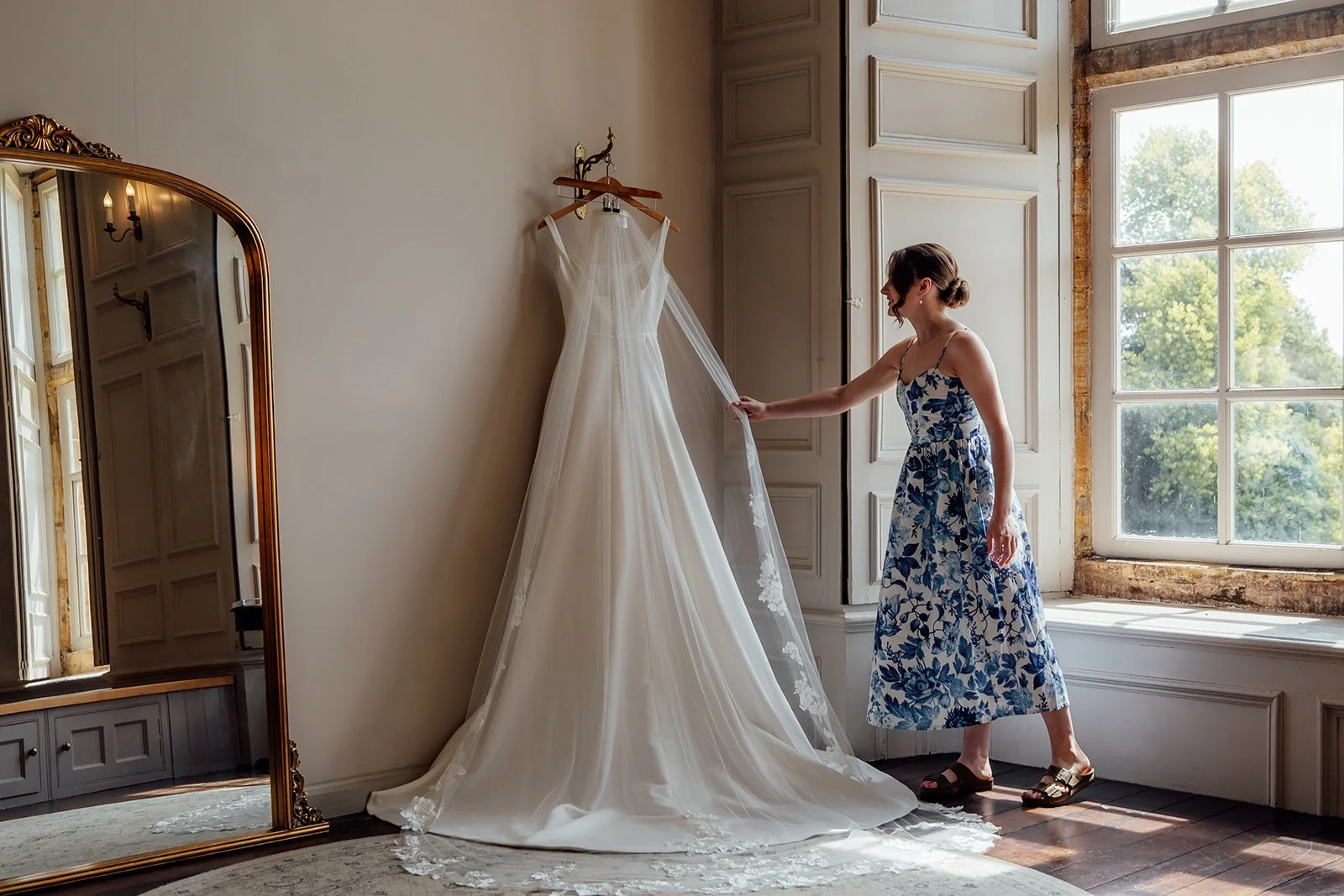 Rachel’s wedding dress hangs beside a tall window as a woman gently adjusts the veil in a bright room at Brympton House in Somerset, photographed by Just James Weddings.