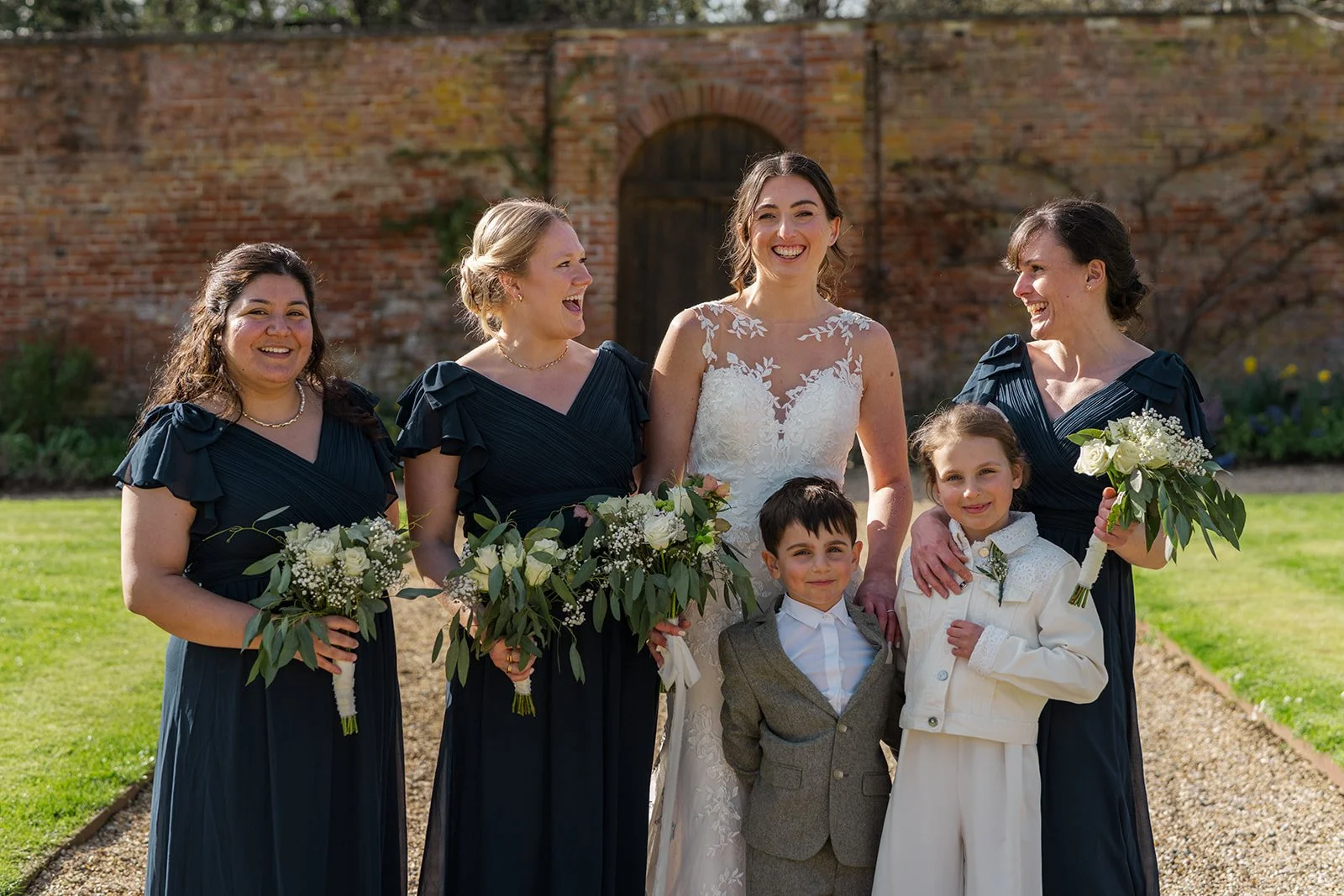 Anna stands with her bridesmaids and two young attendants at Elmhay Park in Somerset, UK, smiling in sunshine with bouquets in hand before the brick garden wall. A bright, joyful group portrait from Anna & Chris’s wedding day in spring light.