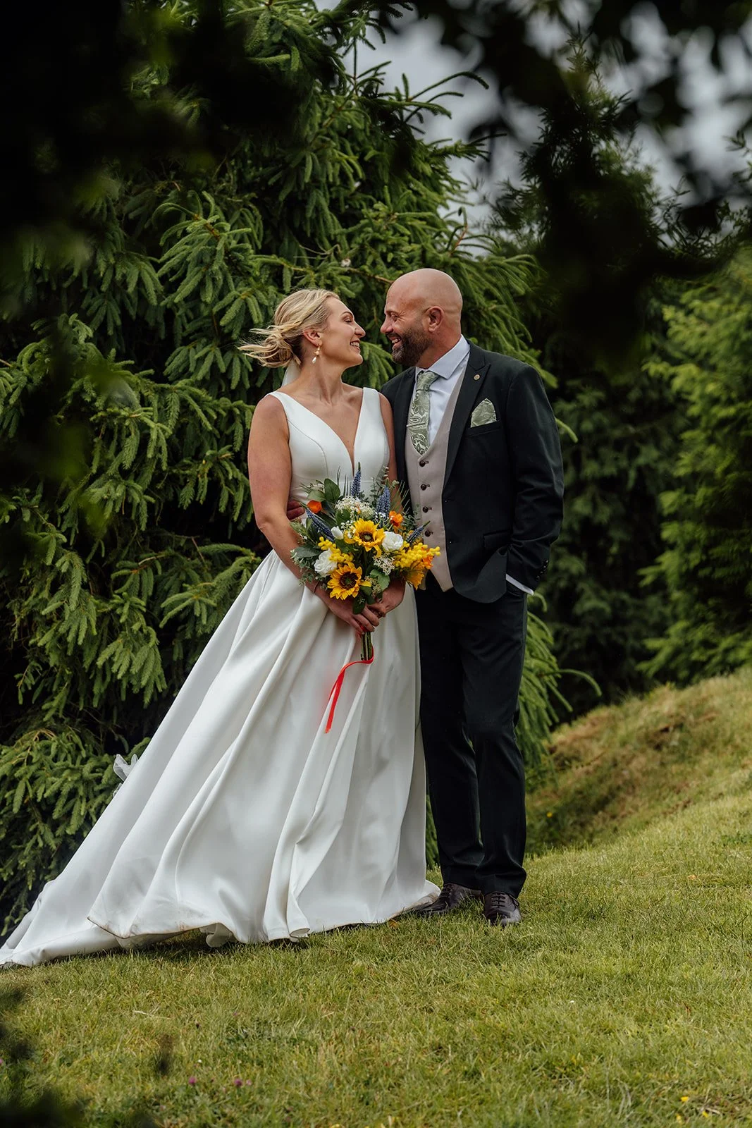 Romantic couple portrait of Hannah & Ben at Grays Barn in Devon, UK, standing among evergreen trees with her sunflower bouquet and his dark suit, captured naturally with soft countryside atmosphere by Just James Weddings.