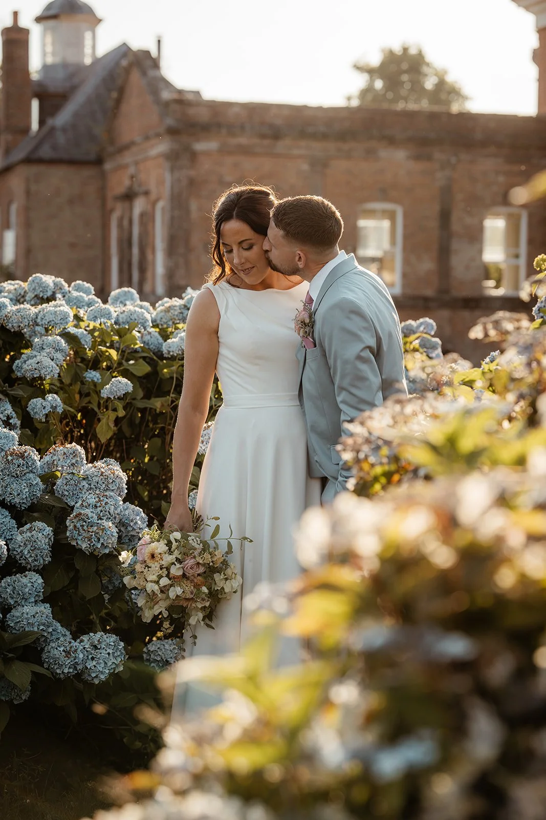 Andy kisses Michelle beside blue hydrangeas in the gardens of Crowcombe Court in Somerset, UK, during a romantic golden-hour wedding portrait.