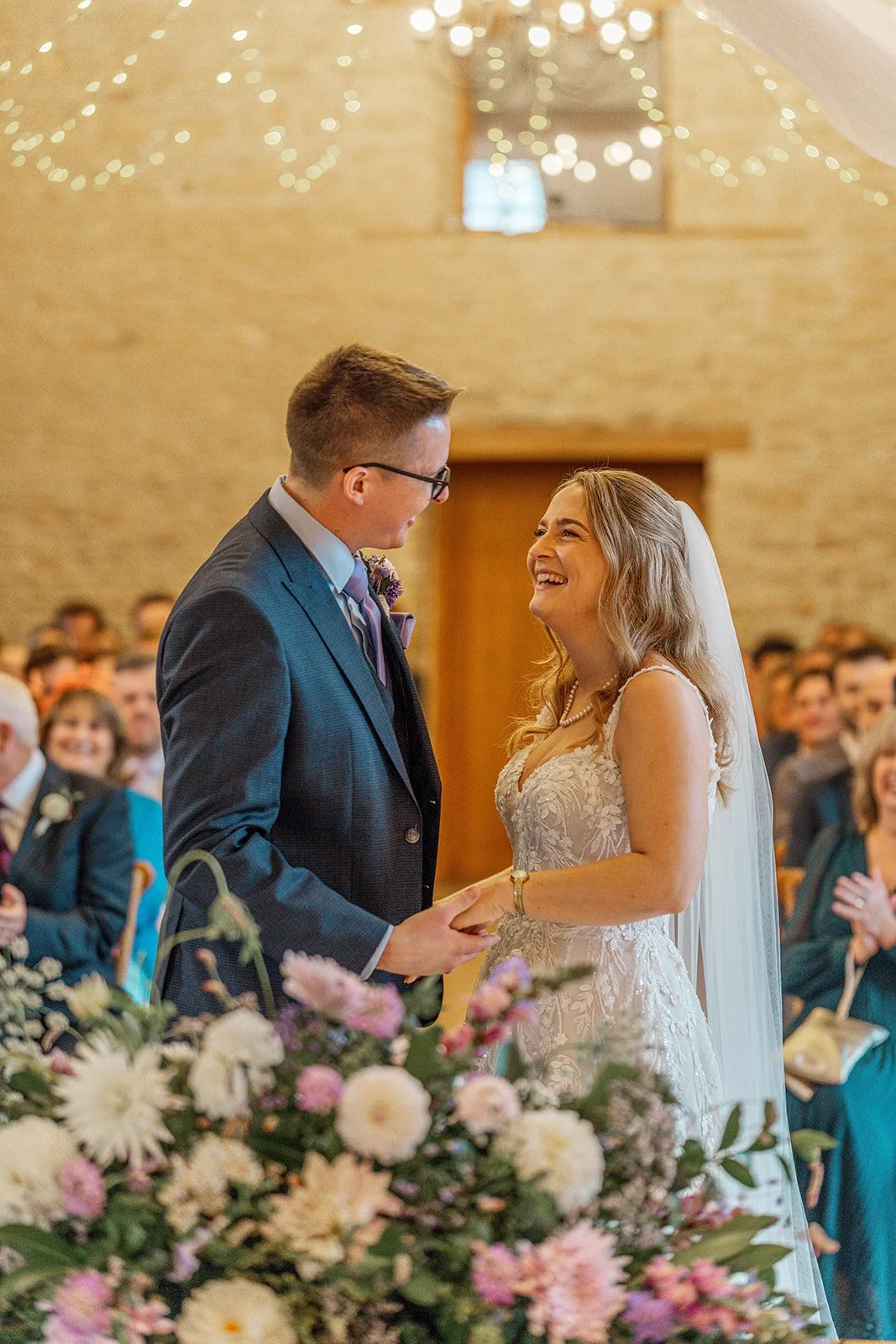 Bride Izzy and groom Andy laugh together during their wedding ceremony, standing before a floral arrangement of pastel blooms inside the warmly lit stone barn at The Kingscote Barn in the Cotswolds, UK, with guests smiling in the background.
