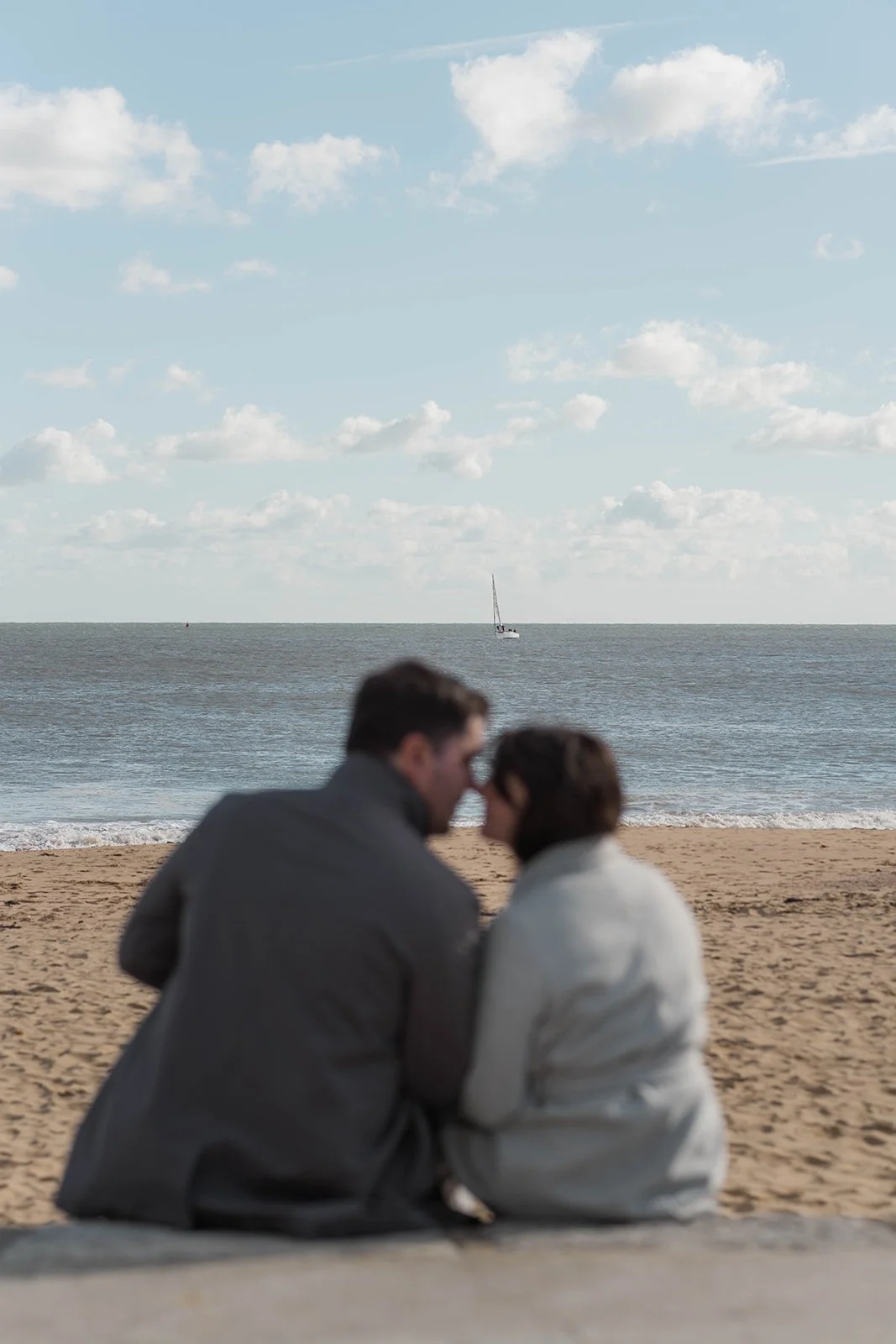 Valentina & Nicky sit together during their engagement shoot by the beach, softly out of focus in the foreground while the sea, pale sand and a lone sailboat fill the frame. The airy composition creates a quiet, intimate and calm coastal portrait.
