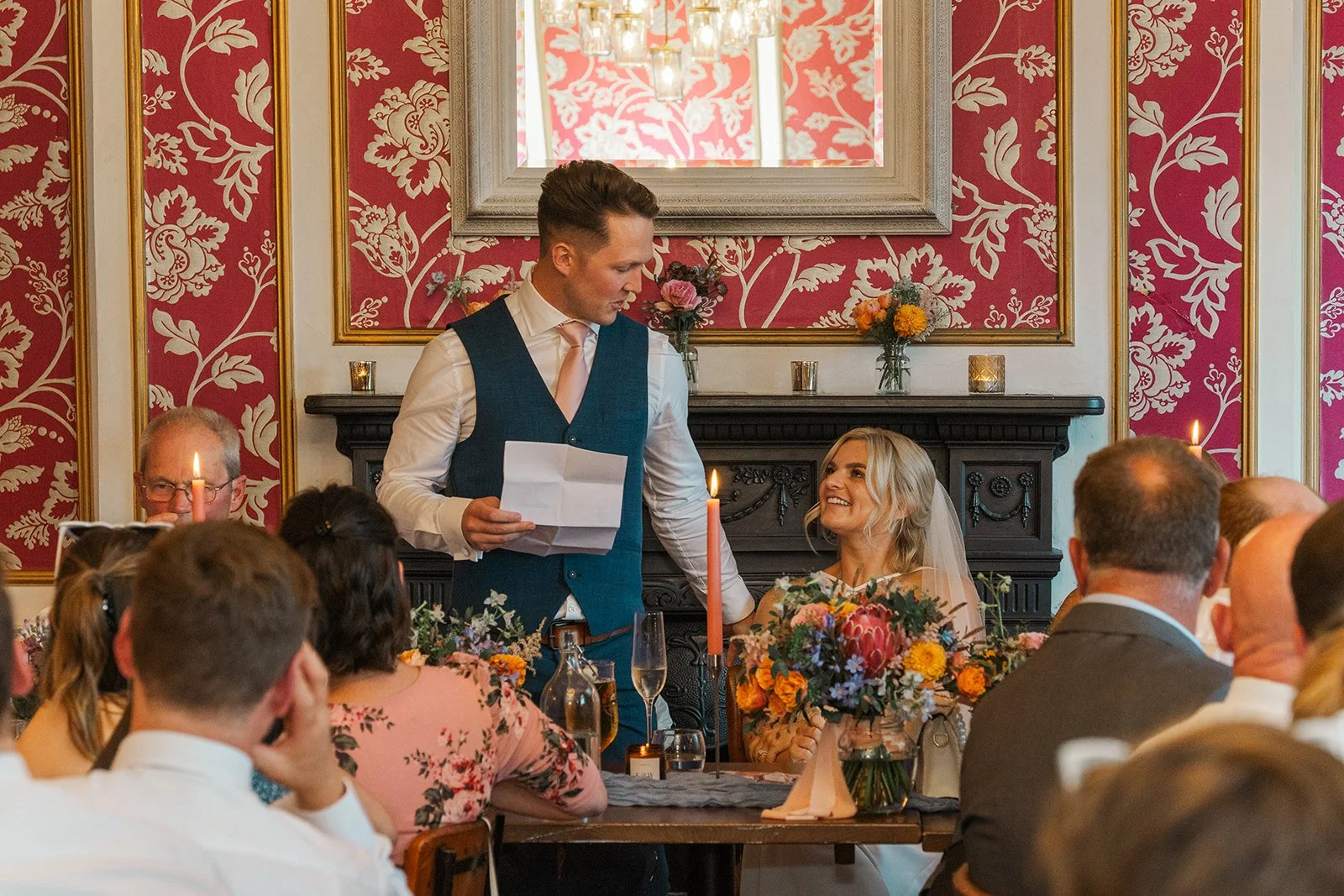 Will gives a wedding speech beside Kristy during their reception at No. 4 Clifton Village in Bristol, UK, surrounded by guests, colourful flowers and the venue’s red patterned décor.