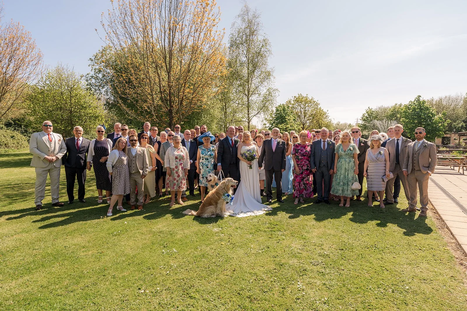 Debbie and Nigel stand with their wedding guests and golden retrievers on the lawn at Yarlington Barn in Somerset, capturing a relaxed group portrait in the sunshine after their countryside wedding ceremony.