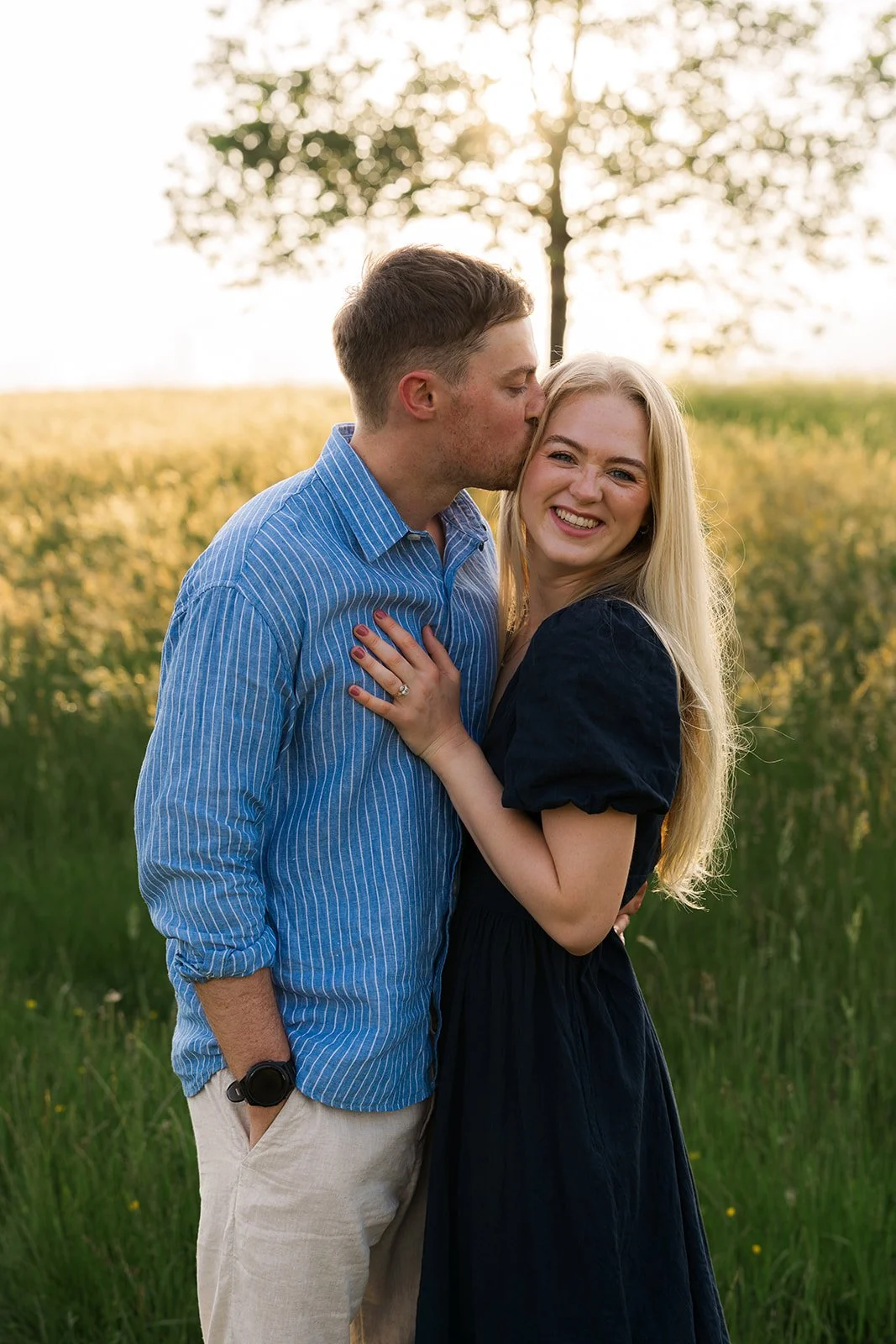Caitlin smiling as Kevin kisses her forehead in a sunny Somerset field outdoors, her engagement ring visible as they stand close together in soft golden evening light during their relaxed countryside engagement photoshoot by Just James Weddings.