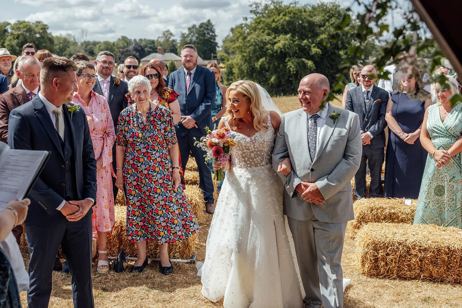 Elise arriving at the outdoor ceremony with her father as Tom smiles at her, surrounded by guests at Bowerchalke Barn in Wiltshire, photographed by Just James Weddings.