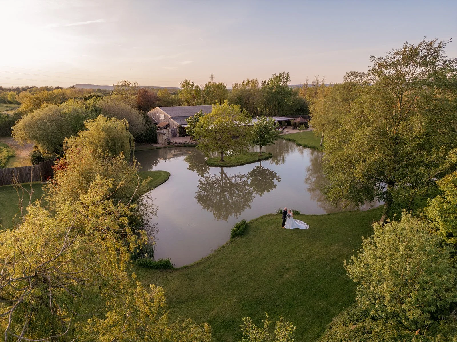 Debbie and Nigel stand beside the lake at Yarlington Barn in Somerset during golden hour, captured from above with the barn, trees and water reflections framing their peaceful countryside wedding portraits.