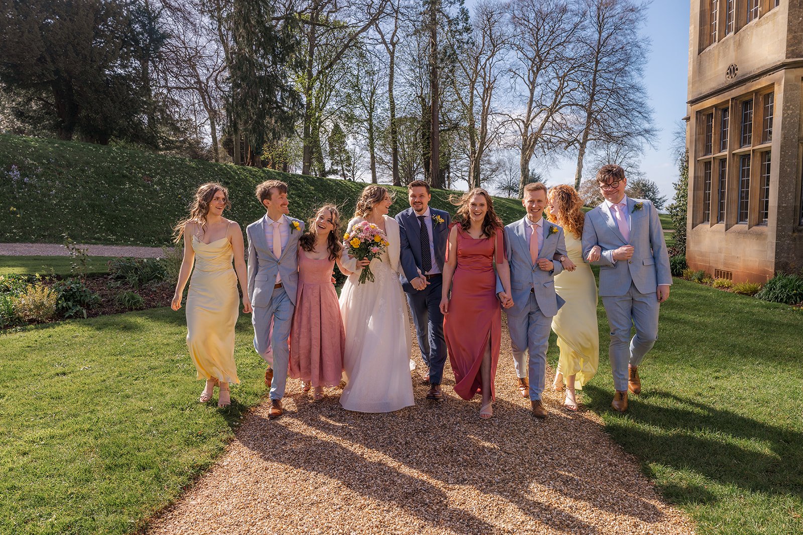 Jessie and Thomas walk with their wedding party through the gardens at Coombe Lodge in Somerset, UK, surrounded by friends in colourful dresses and suits.