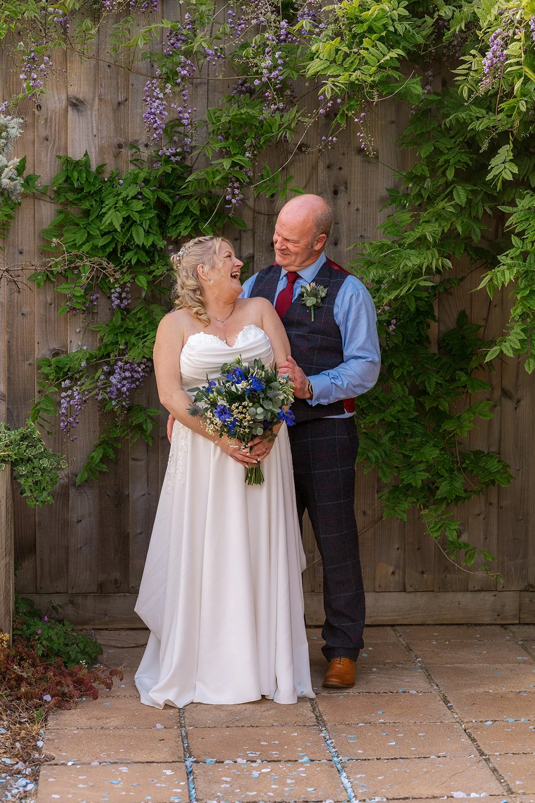 Debbie and Nigel laugh together beneath purple flowers at Yarlington Barn in Somerset, standing against the garden fence with Debbie’s blue bouquet for a relaxed couple portrait after their sunny countryside wedding.