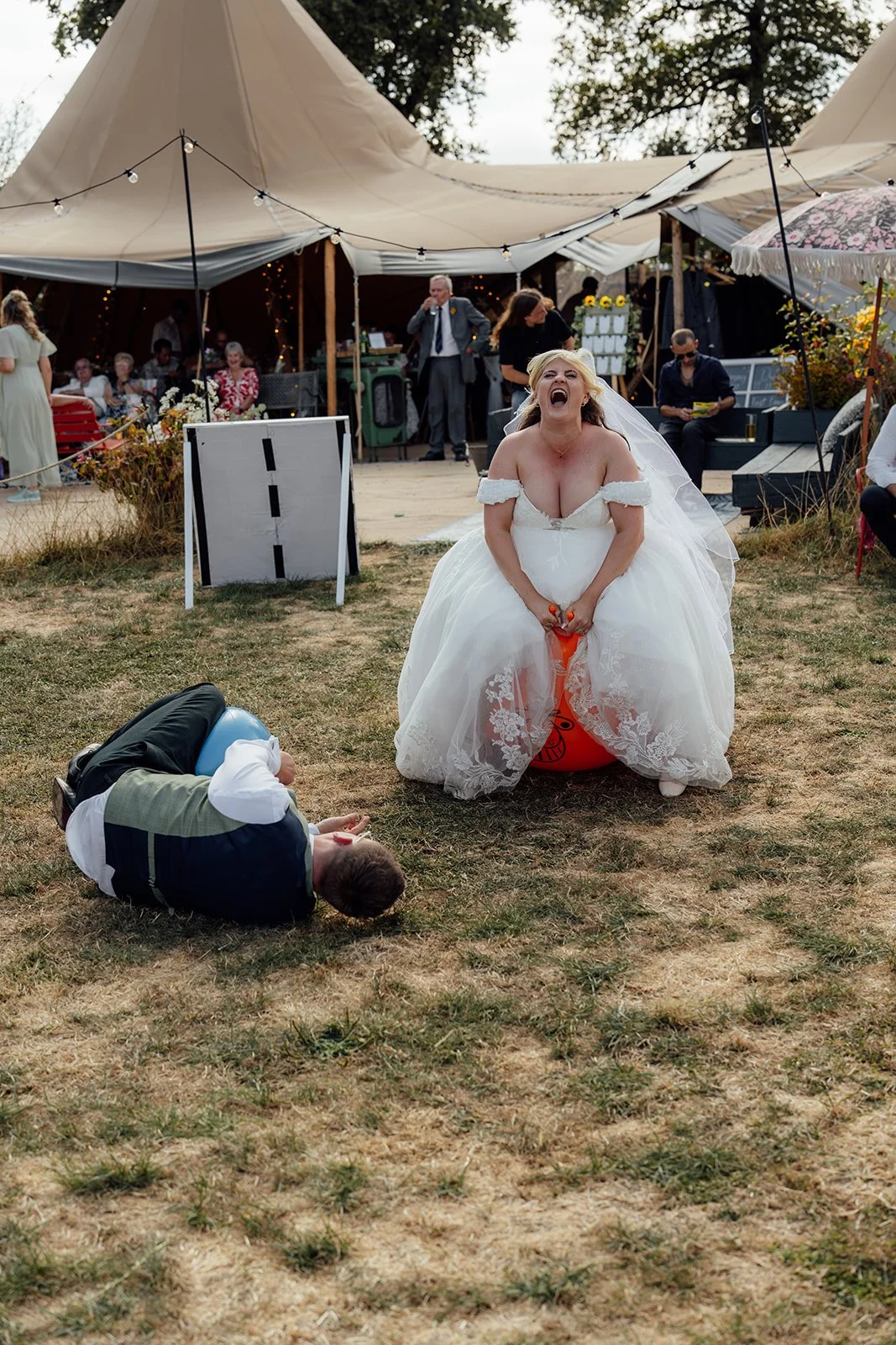 Lucy laughing while sitting on a space hopper as Matthew falls beside her during a playful outdoor wedding moment at Curated Canvas at Noah’s Pond in Somerset, photographed by Just James Weddings.