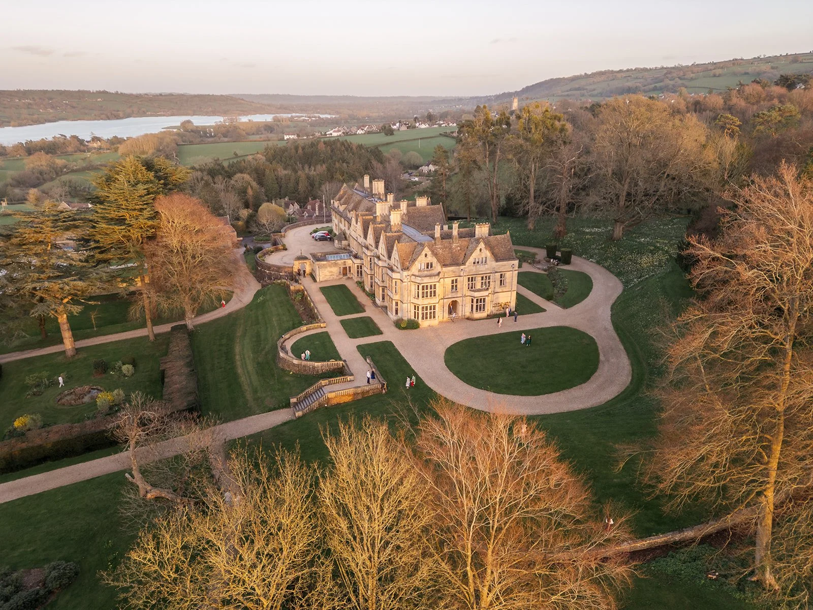 Aerial drone view of Coombe Lodge in Somerset, UK, surrounded by landscaped grounds, woodland and sweeping countryside in the warm evening light.