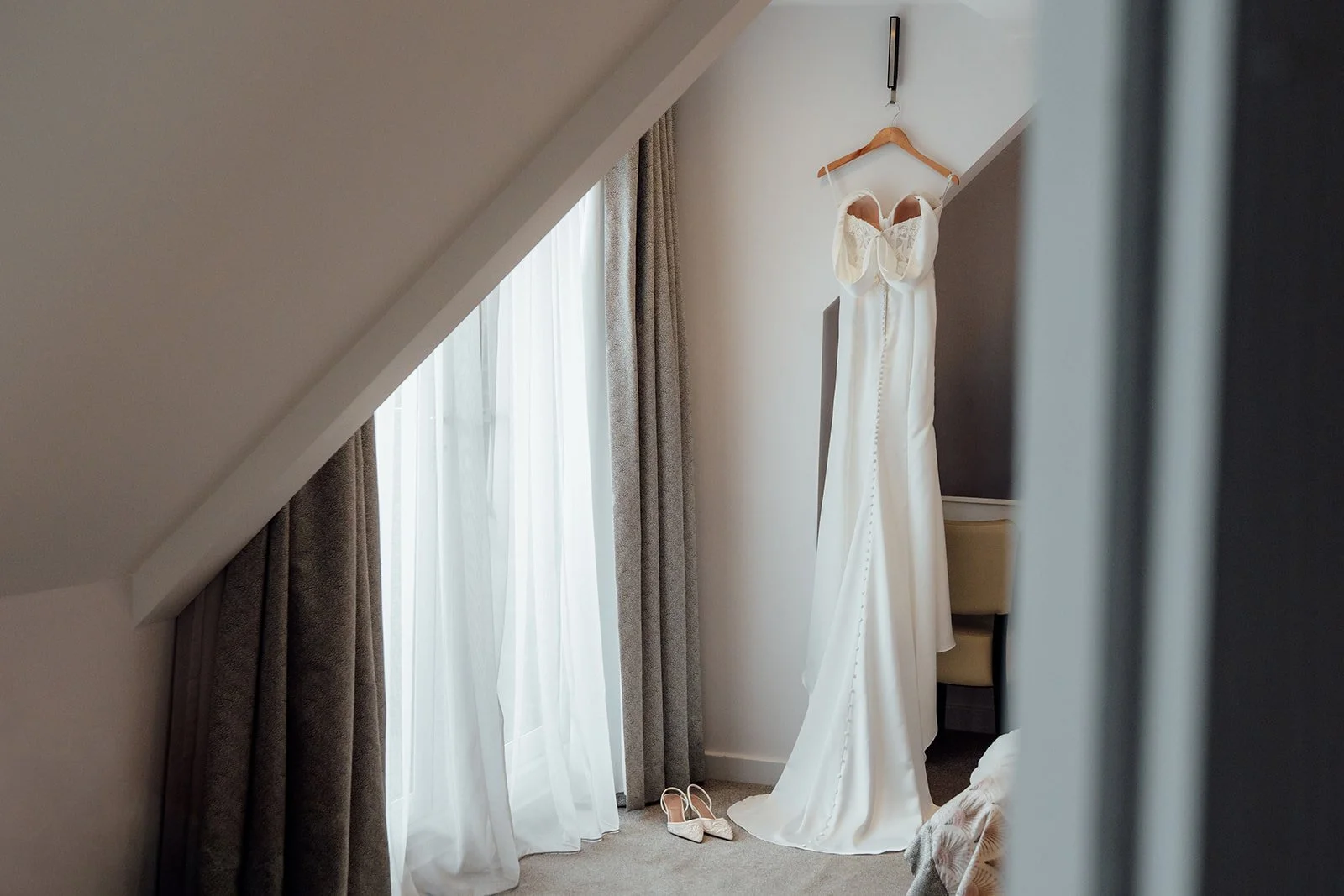 Amy’s fitted wedding dress hangs beside a bright window with her bridal shoes below, photographed during morning preparations at Sandy Cove Hotel in North Devon, UK.