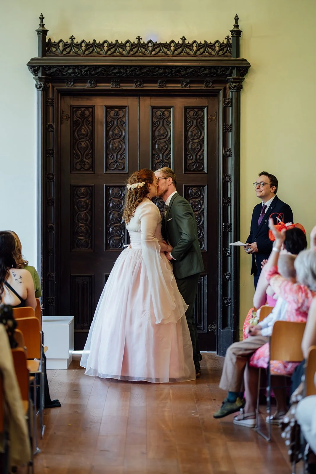 Charlotte & Jack share their first kiss during their Bishop’s Palace wedding ceremony in Somerset, UK, standing before carved wooden doors as guests watch from the aisle and the registrar smiles beside them inside the historic ceremony room.