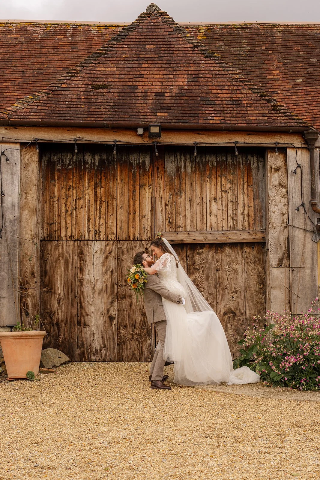 Maisie & Andrew sharing a playful kiss outside the rustic barn at Stockbridge Farm Barn in Dorset, photographed by Just James Weddings for fun autumn wedding portraits.